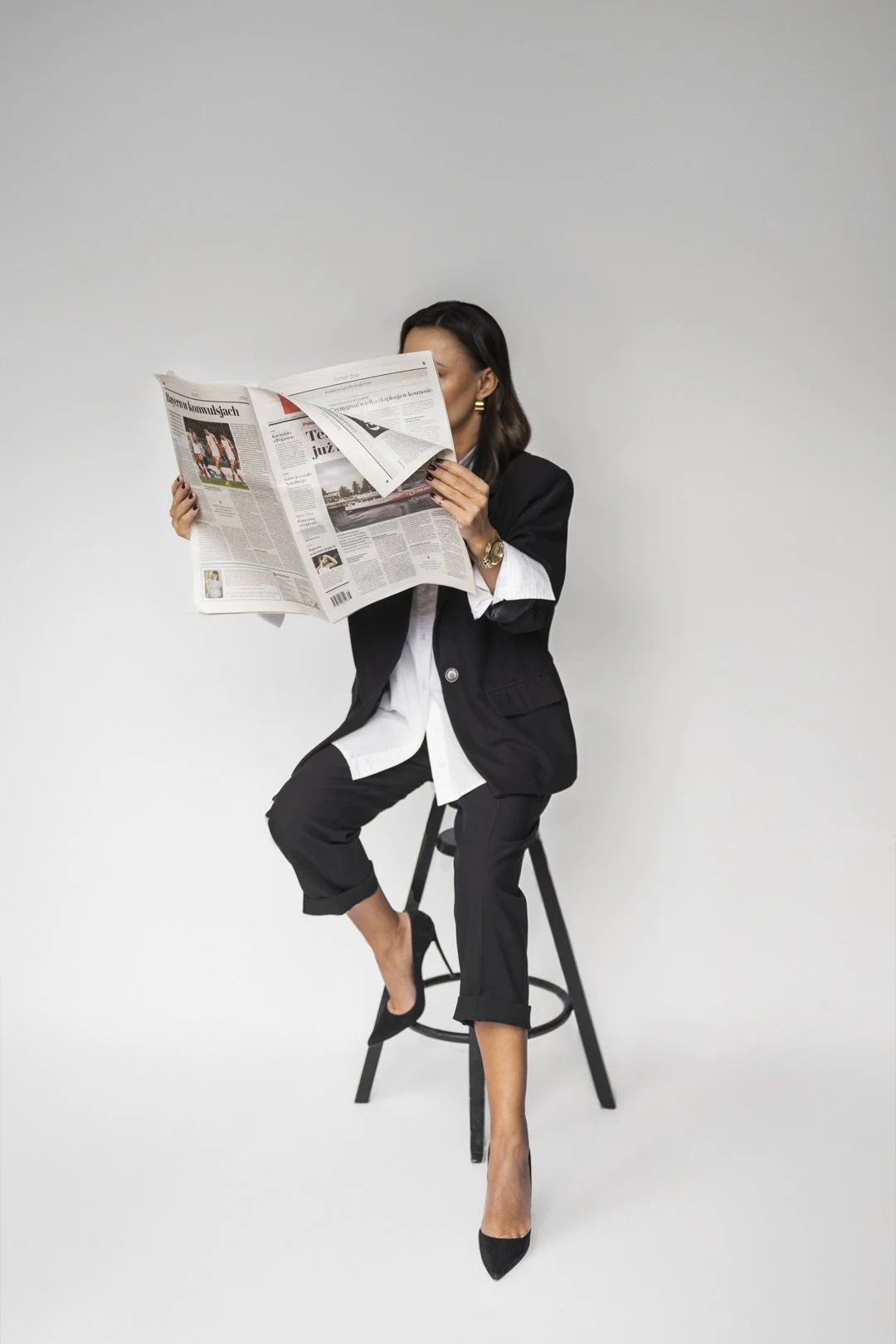 A woman dressed in formal black and white attire sitting on a black stool, reading a newspaper against a plain white background.