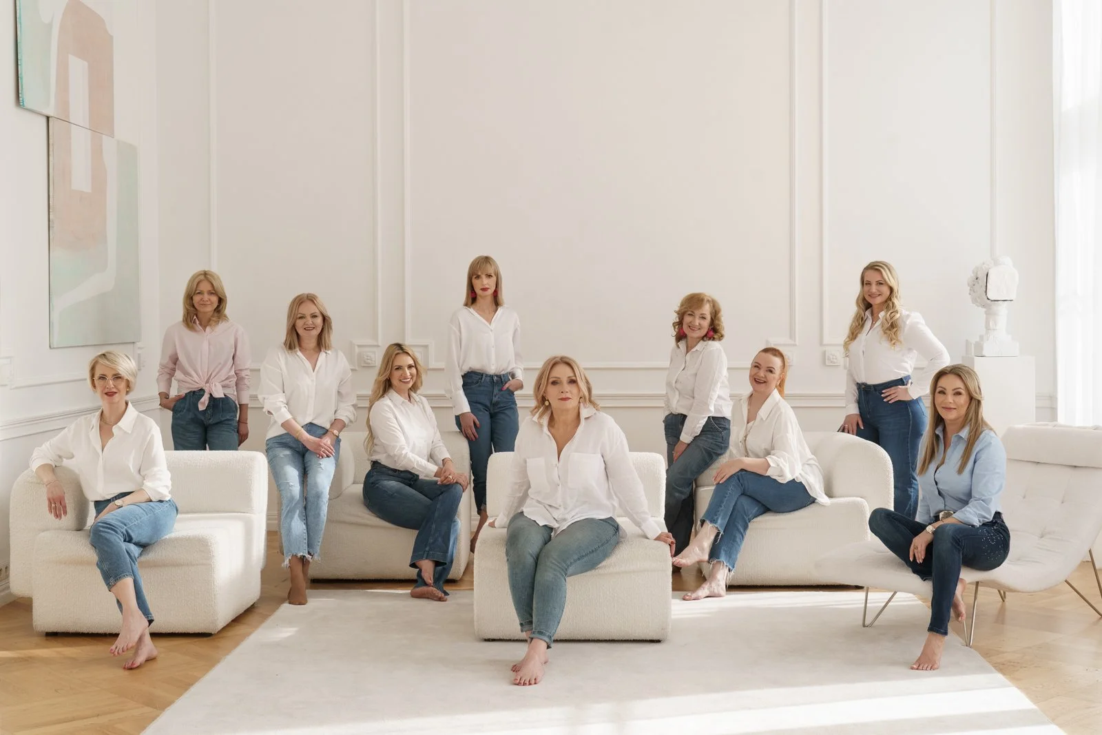 A group of eleven women in white and blue shirts and jeans posing in a bright, modern living room with white walls, artwork, and contemporary furniture.