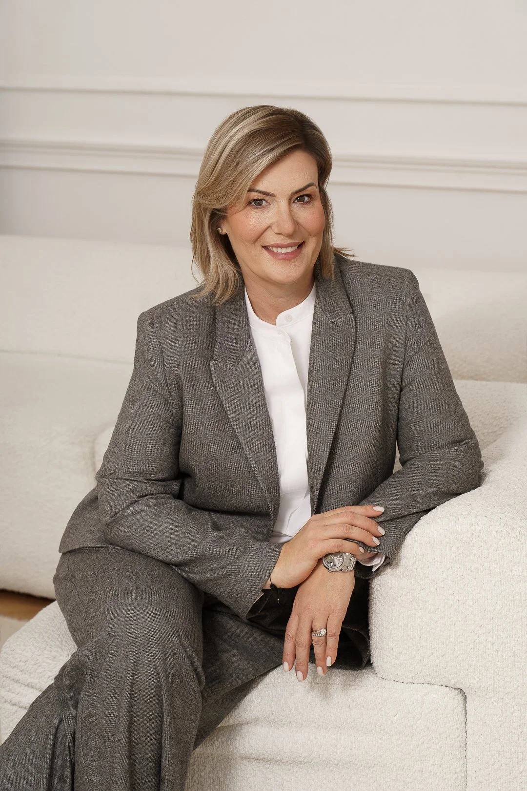 A smiling woman with blonde hair dressed in a gray suit and white shirt, sitting on a white sofa.