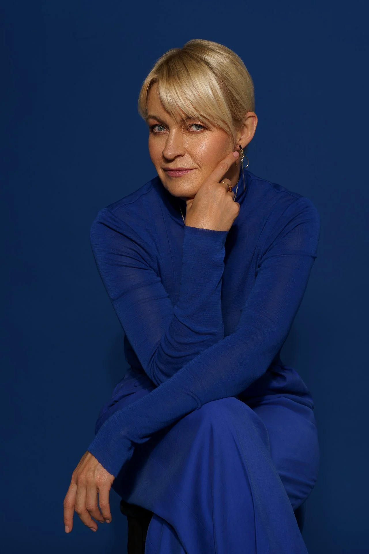 A woman with short blonde hair wearing a blue long-sleeve shirt and pants, sitting against a dark blue background, with her left hand resting on her chin and her right hand hanging down.