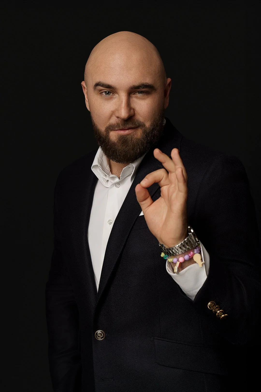 A man with a beard, wearing a white shirt and dark blazer, making an OK gesture with his right hand, standing against a black background.