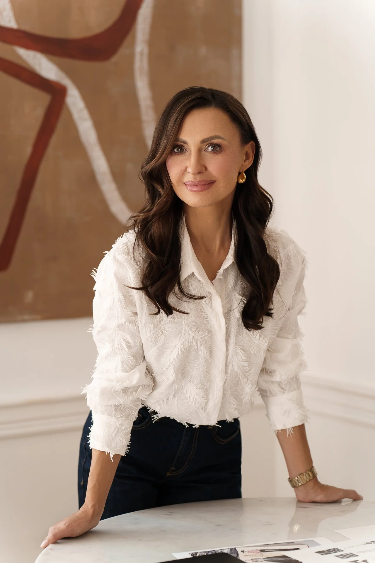 A woman with long dark hair, wearing a white textured shirt and gold jewelry, standing and leaning on a marble table in a well-lit room.