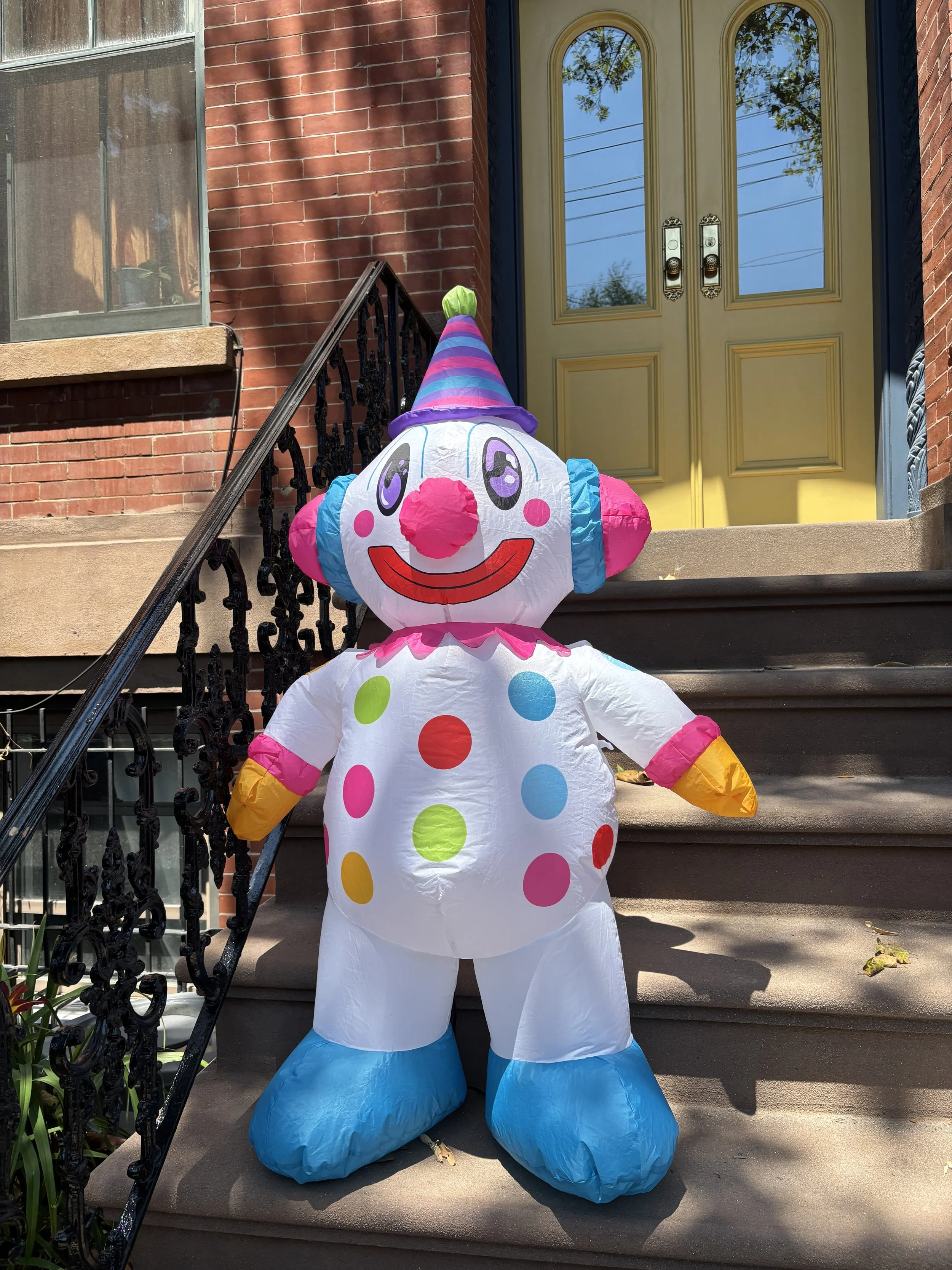 Cutesy is a 4-foot lighted seasonal decorative inflatable. Cutesy stands on the front porch steps. The house has a yellow door and black iron railing. Cutesy glows after dark.