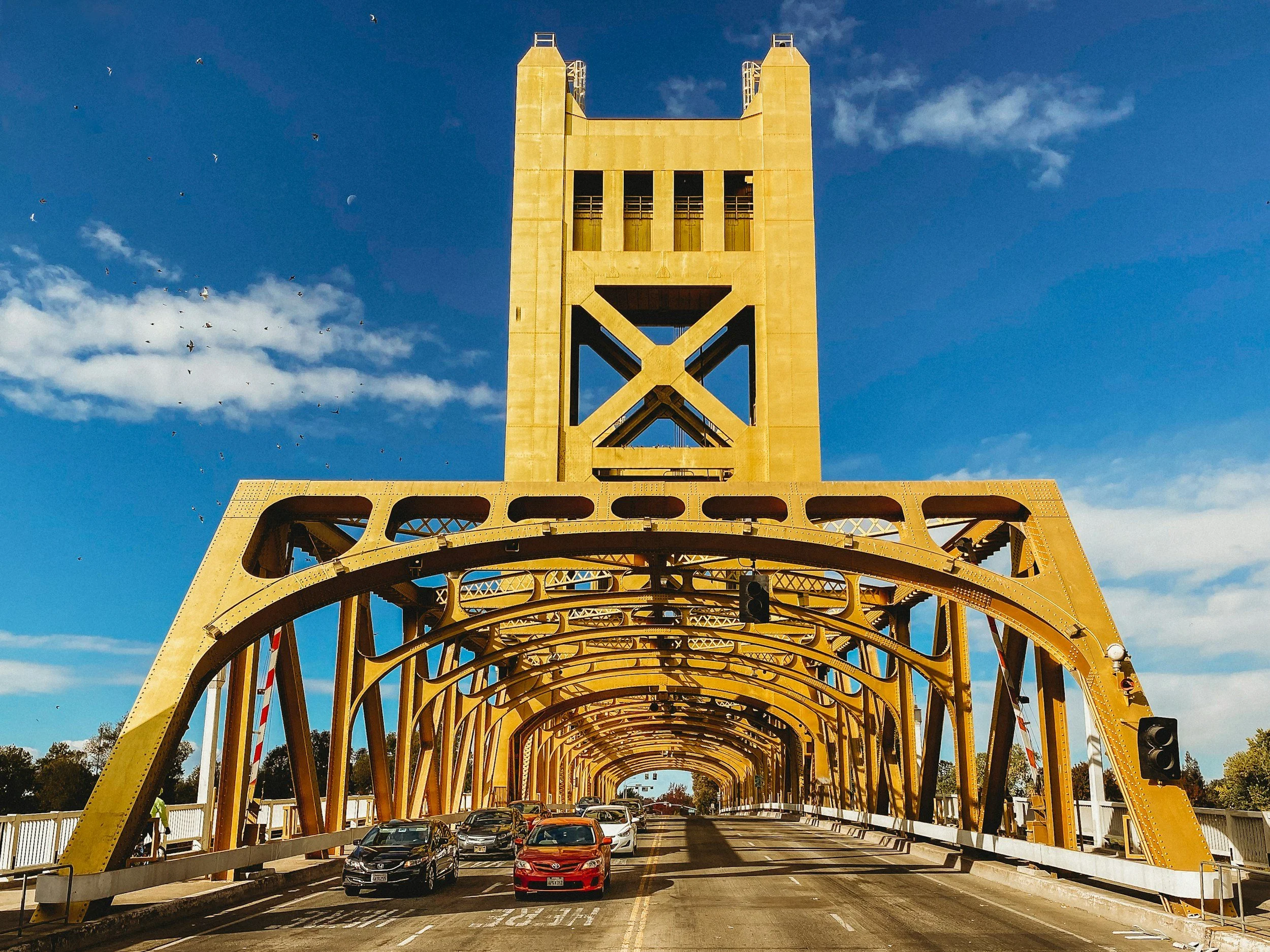 Golden bridge with a tower in the background and cars driving on it, under a blue sky with some clouds and birds.