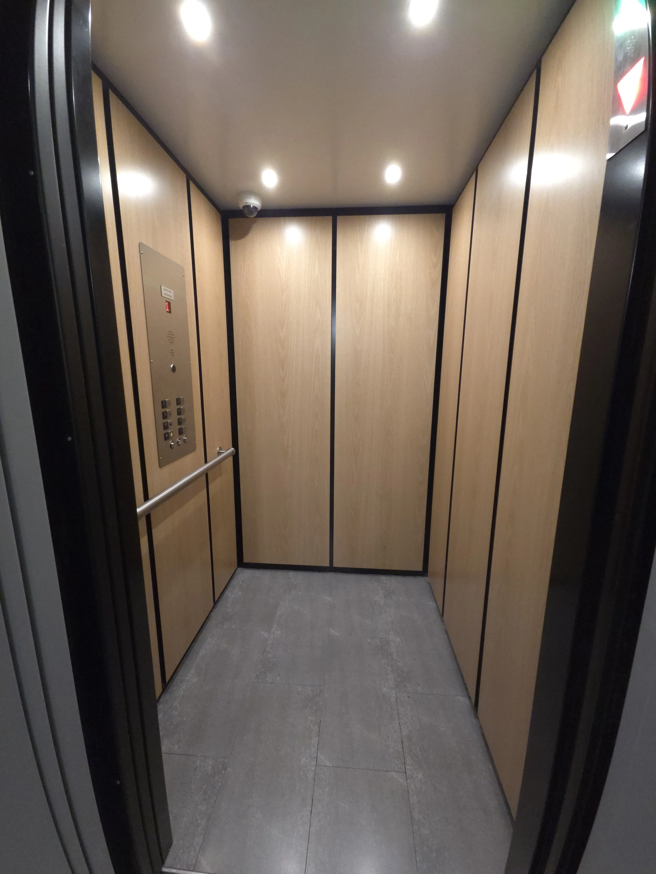 Empty elevator with wood-paneled walls, gray tiled floor, ceiling lights, an elevator control panel on the left wall, and a smoke detector on the ceiling.
