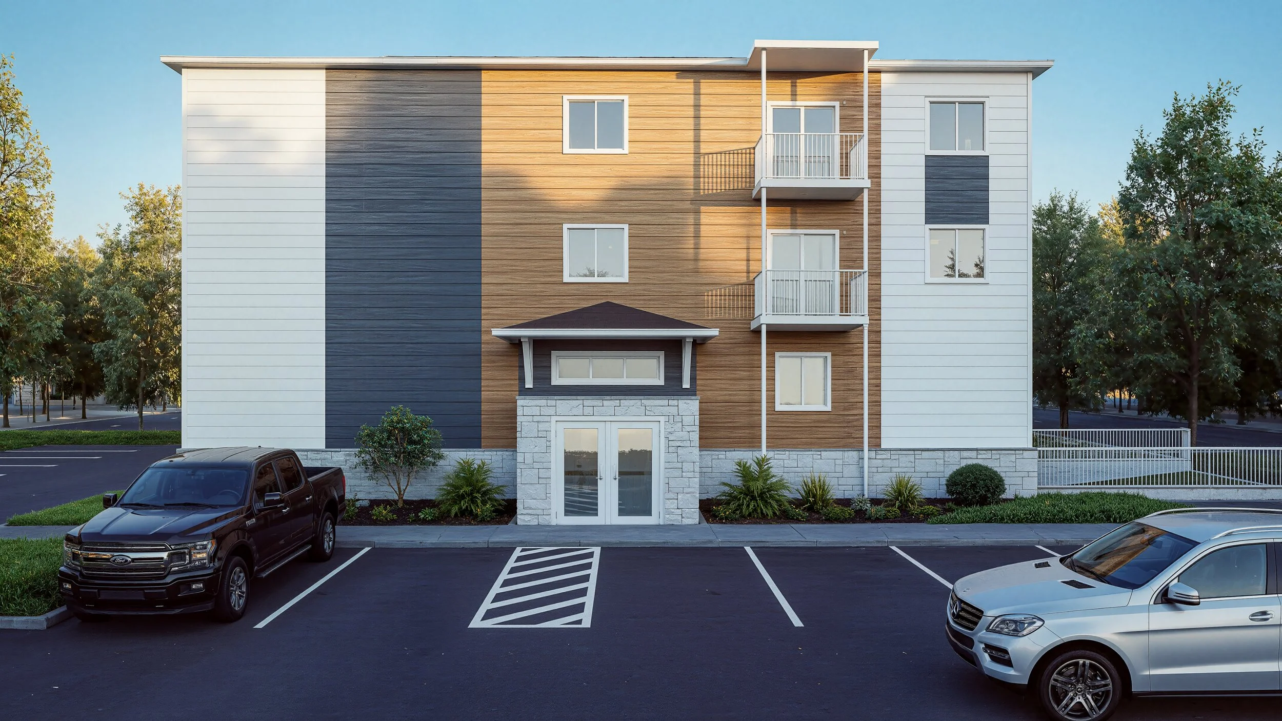 Front view of a multi-story apartment building with a parking lot in the foreground, two cars parked, and trees in the background.