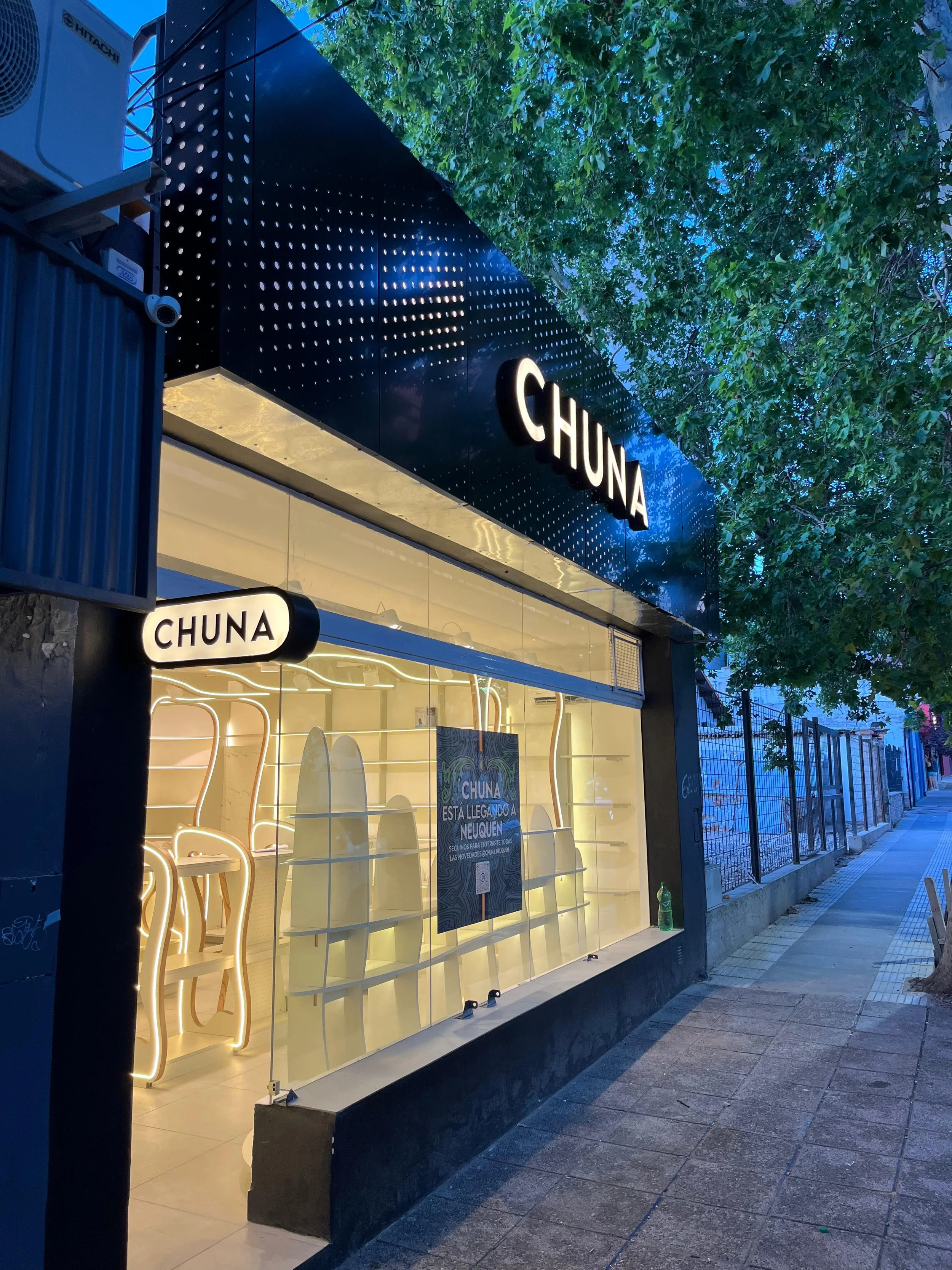 Fachada de una tienda llamada CHUNA con iluminación moderna y escaparate con tablas de surf decorativas, en una calle con árboles y un cielo al atardecer.