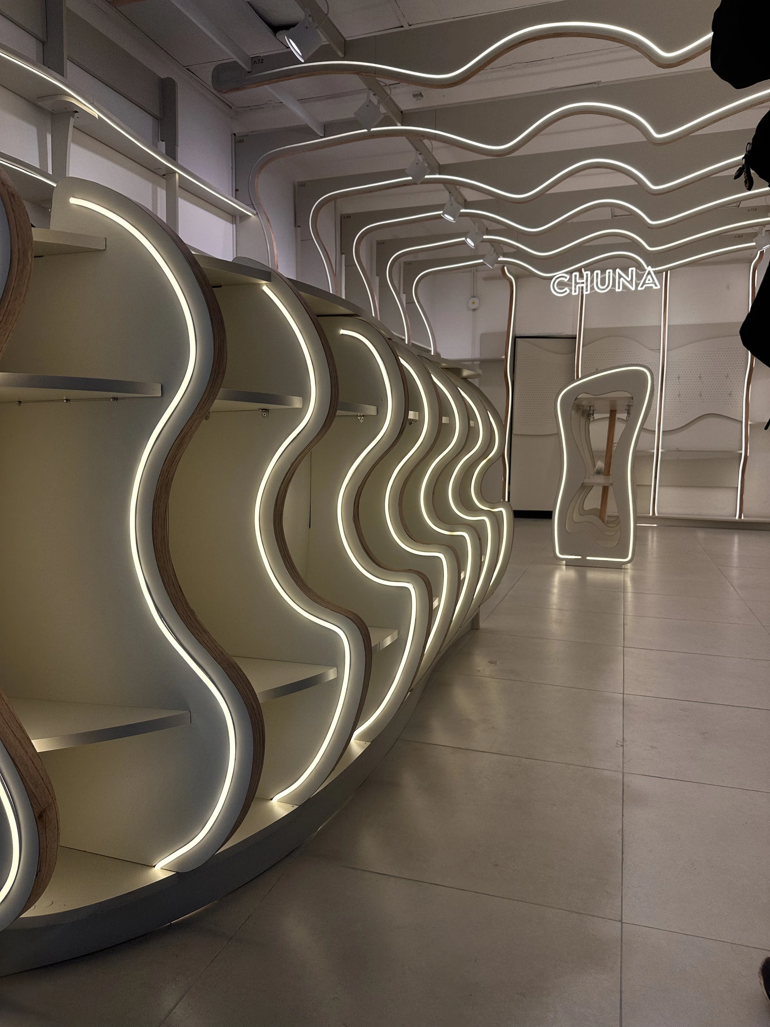 Estanterías con iluminación de neón blanco en forma de ondas, en un espacio moderno con decoración de líneas curvadas y letras de neón que dicen 'CHUNA'.