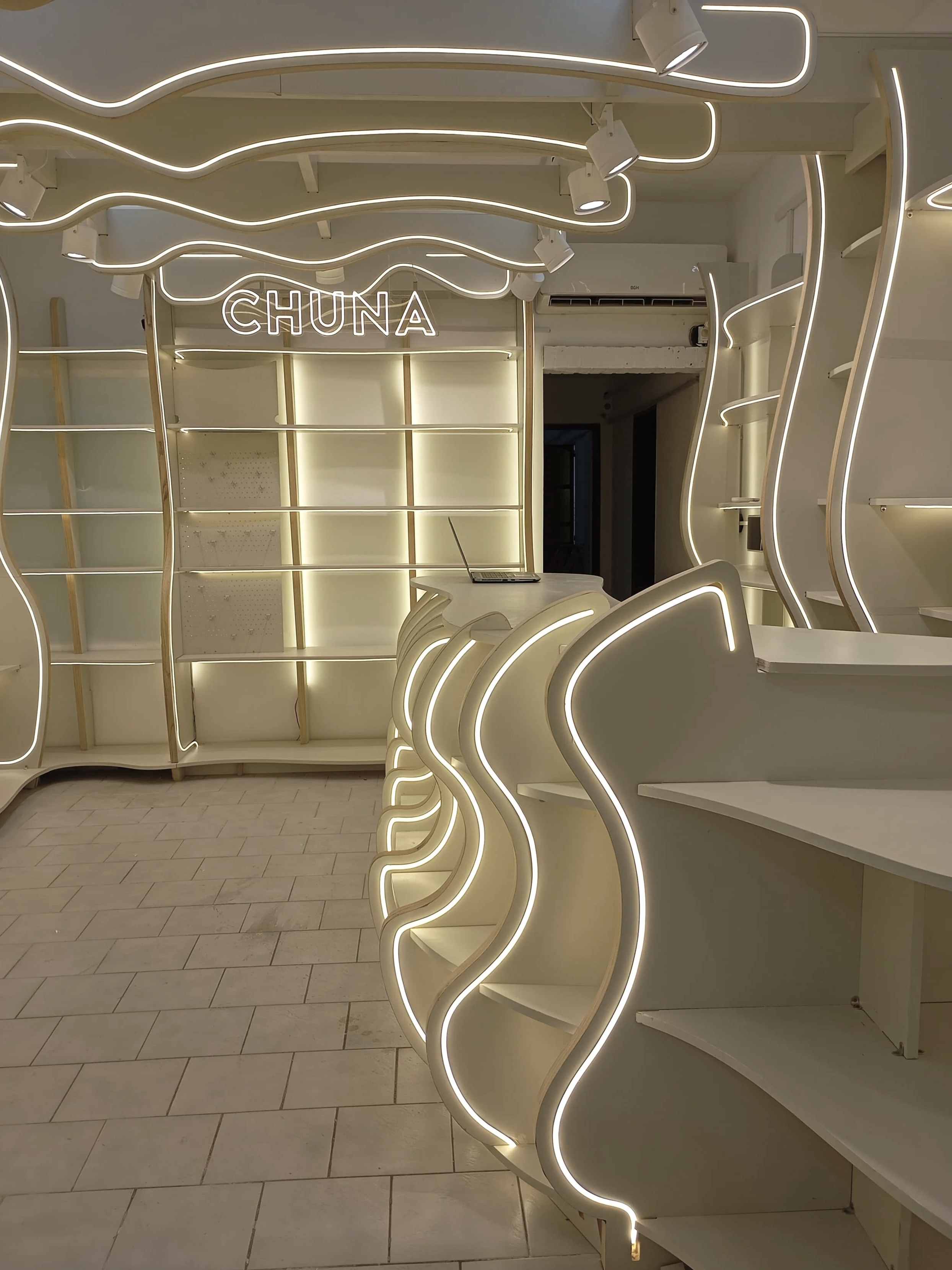 Estanterías blancas con luces LED en un espacio con el cartel 'CHUNA' en la pared y una laptop en una de las mesas.