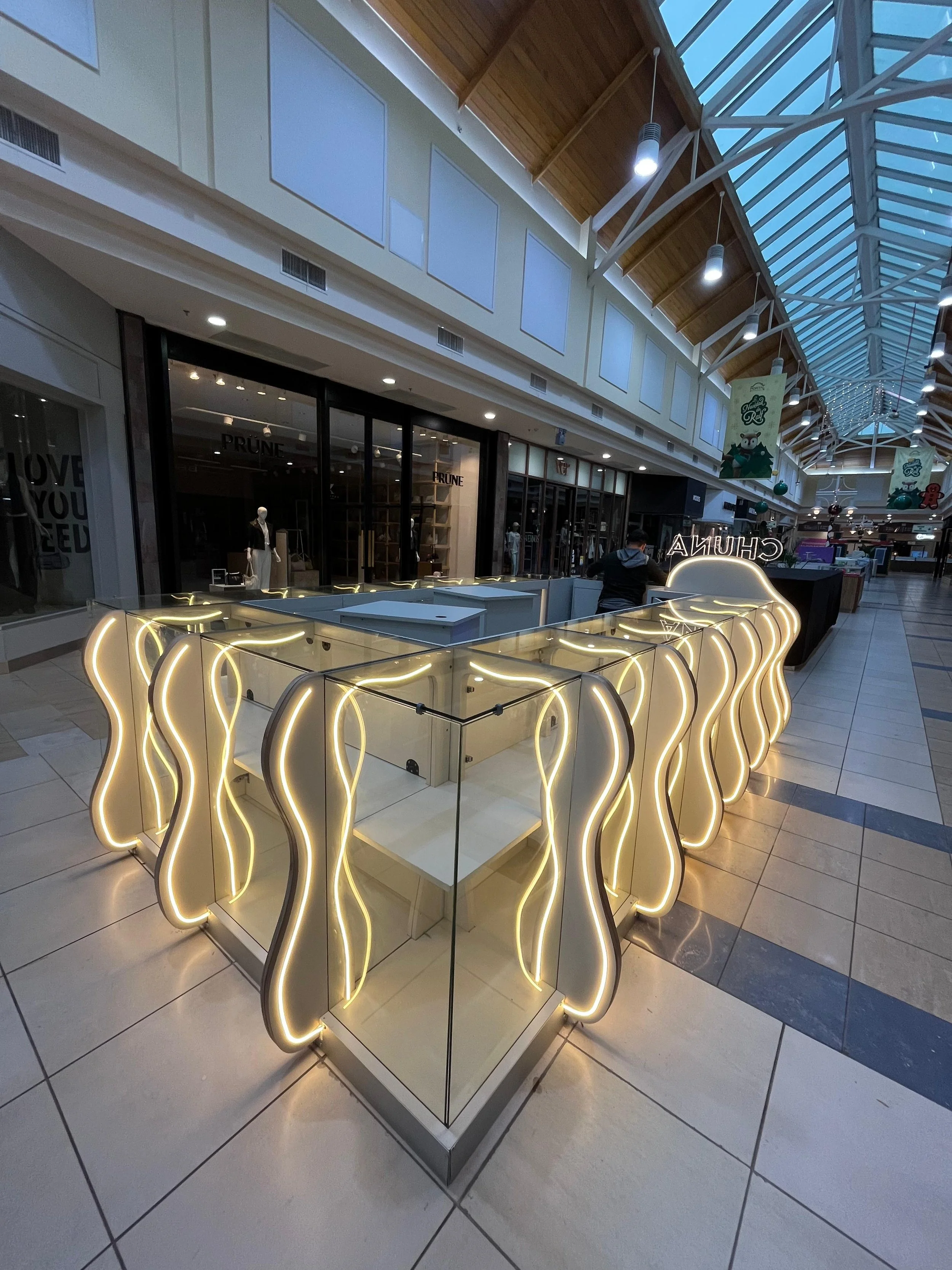 Vitrina de exhibición en un centro comercial con iluminación decorativa en forma de curvas y un fondo de tiendas y pasillo cubierto con techo de vidrio.