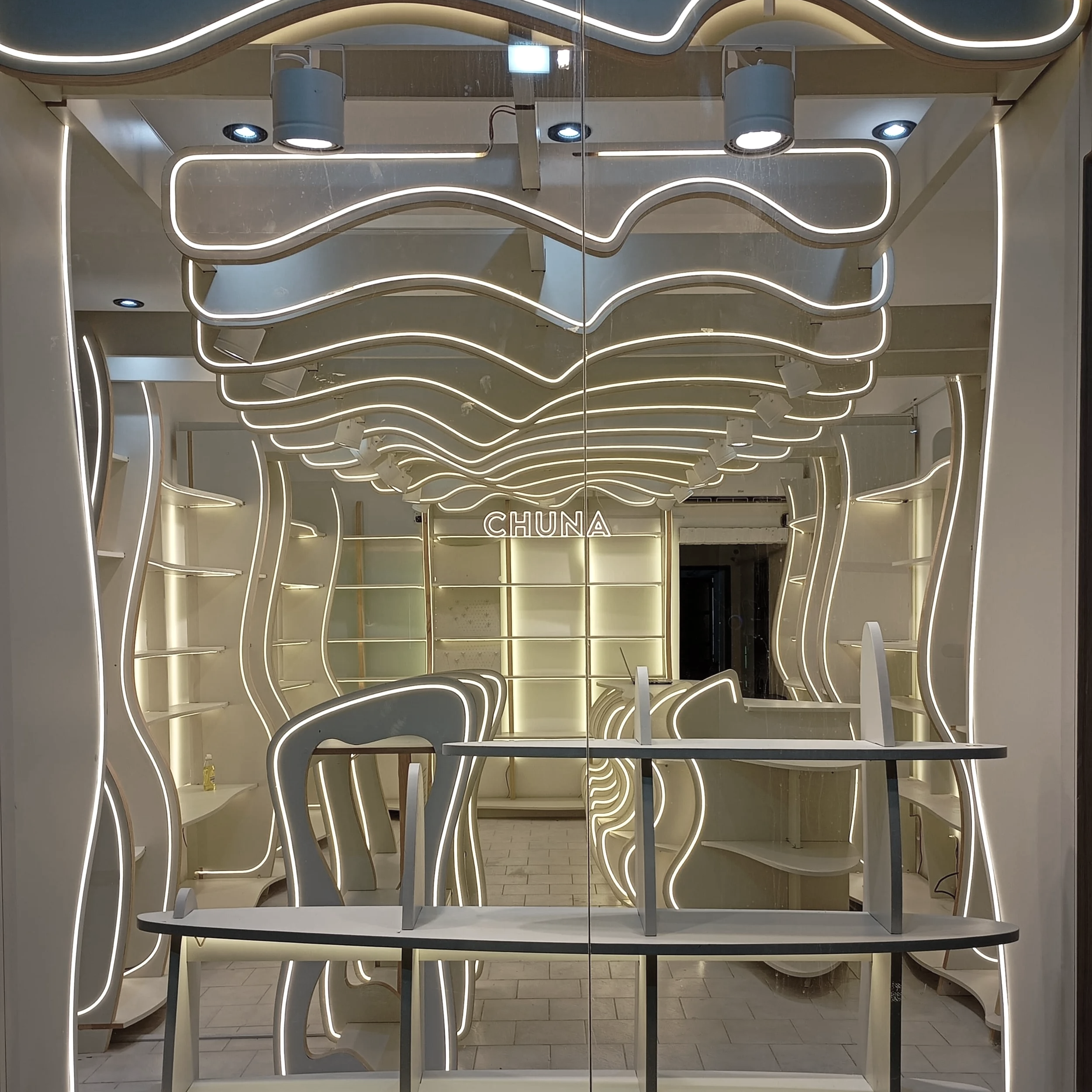 Interior de una tienda con techo y paredes de diseño curvado iluminado con luces LED y una repisa en la parte trasera, con letrero de neón que dice 'CHUNA'.