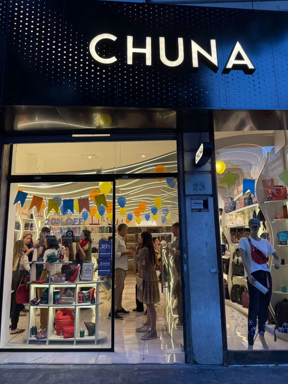 Vista del exterior de una tienda llamada 'Chuna' con muchas personas adentro, decorada con globos de colores y promociones de descuentos.