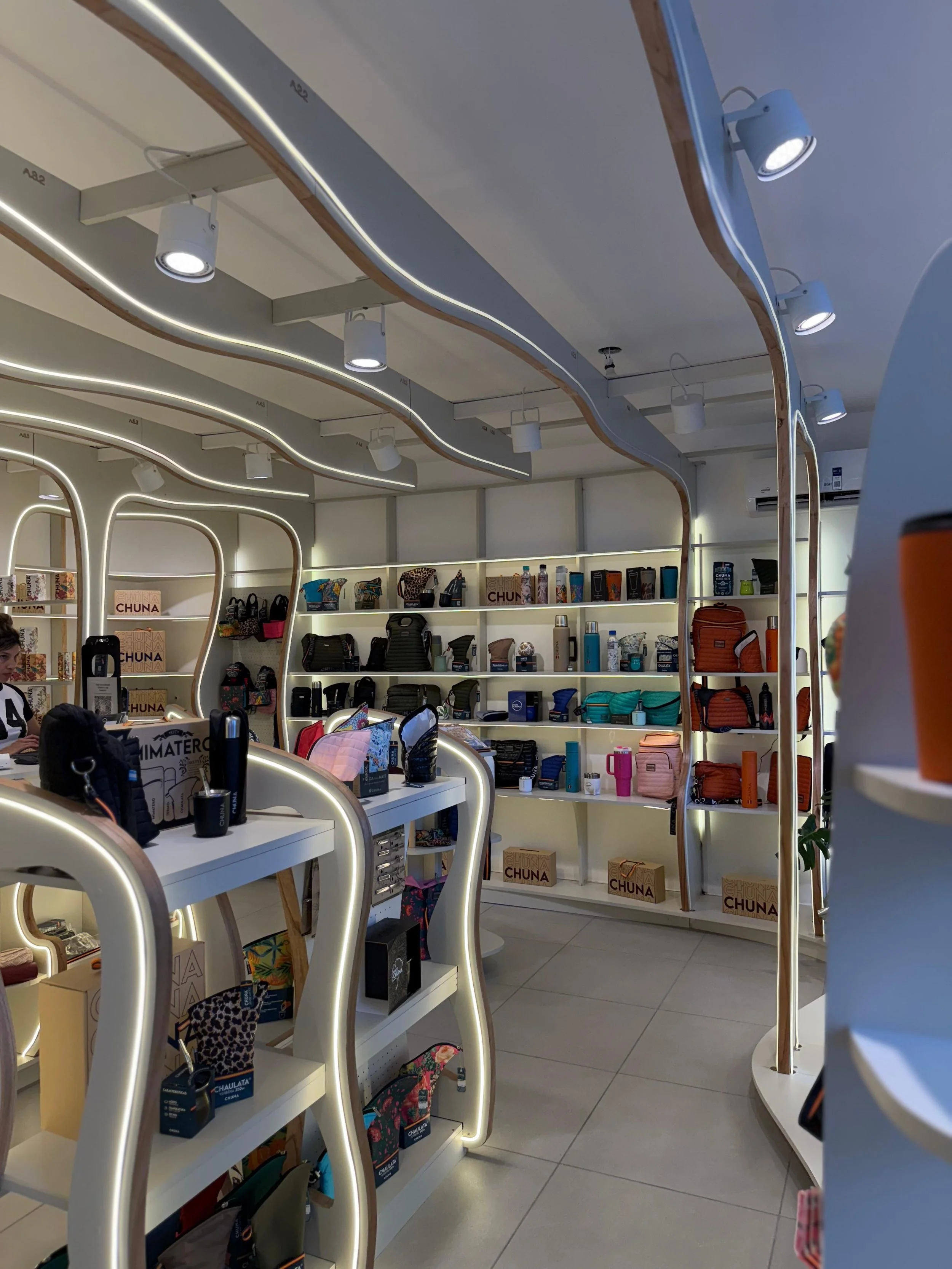 Vista interior de una tienda con estantes llenos de accesorios y productos, incluyendo bolsas, termos, y otros artículos, con iluminación moderna y diseño ordenado.