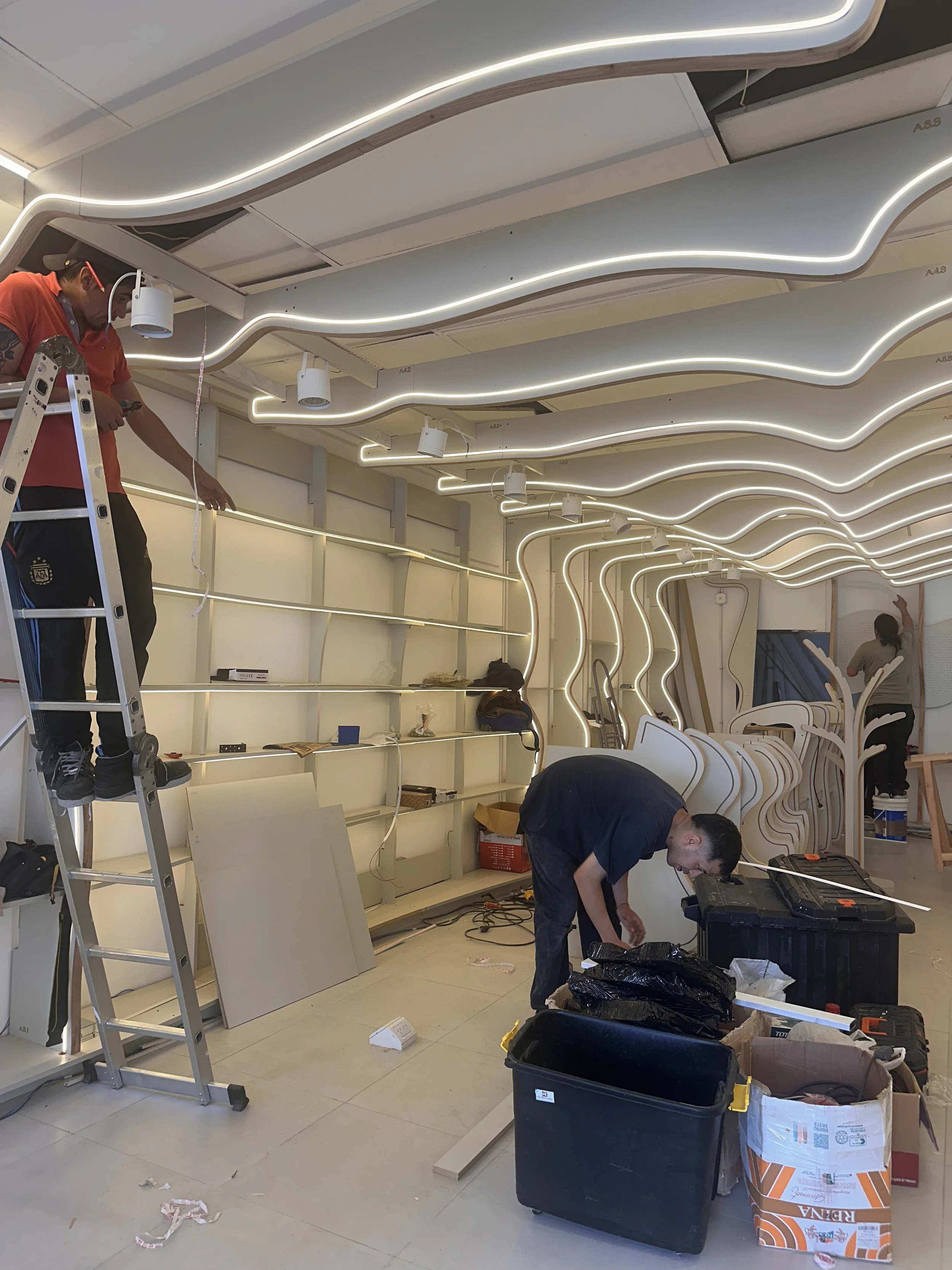 Personas instalando iluminación y mobiliario en una tienda moderna en construcción, con lámparas de neón y estanterías aún en proceso.