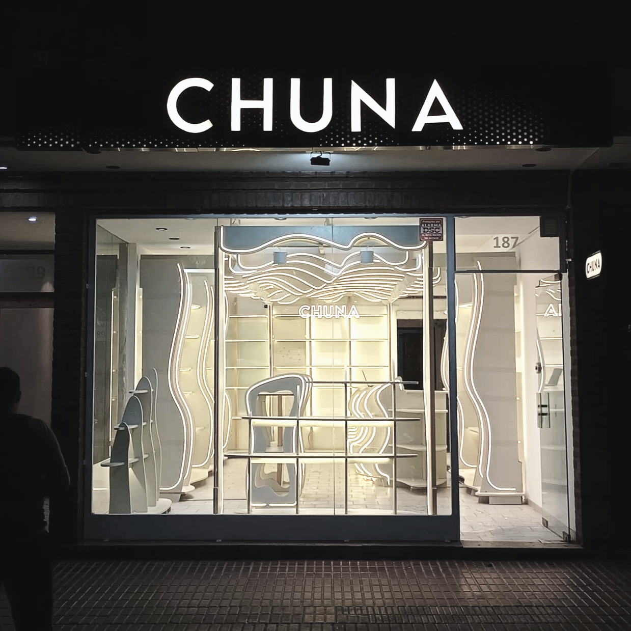 Fachada de una tienda llamada CHUNA, con iluminación de neón y decoraciones modernas en el interior, vista nocturna.