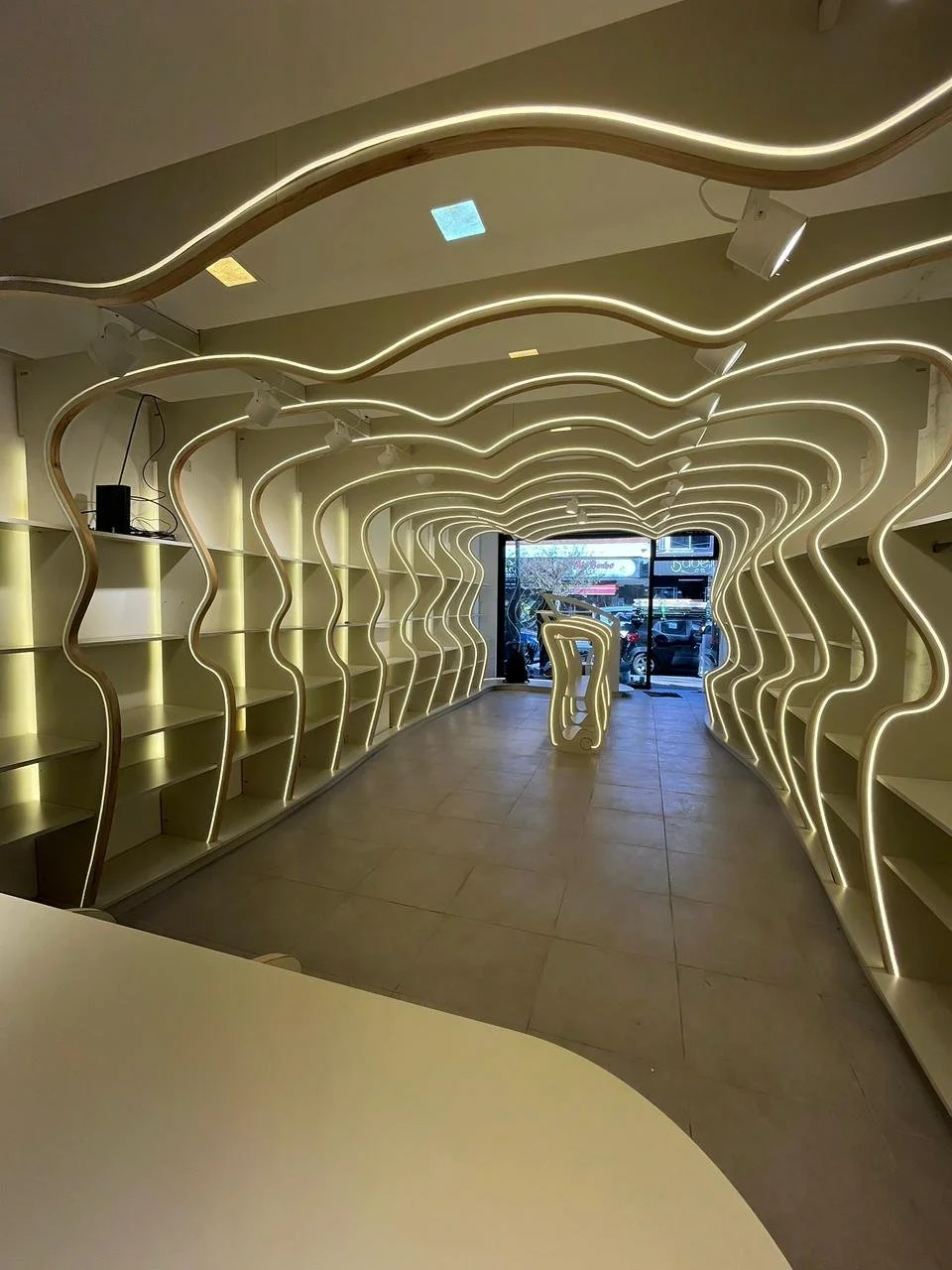 Interior moderno con estanterías y un pasillo con iluminación en forma de ondas, con una fachada de vidrio al fondo.