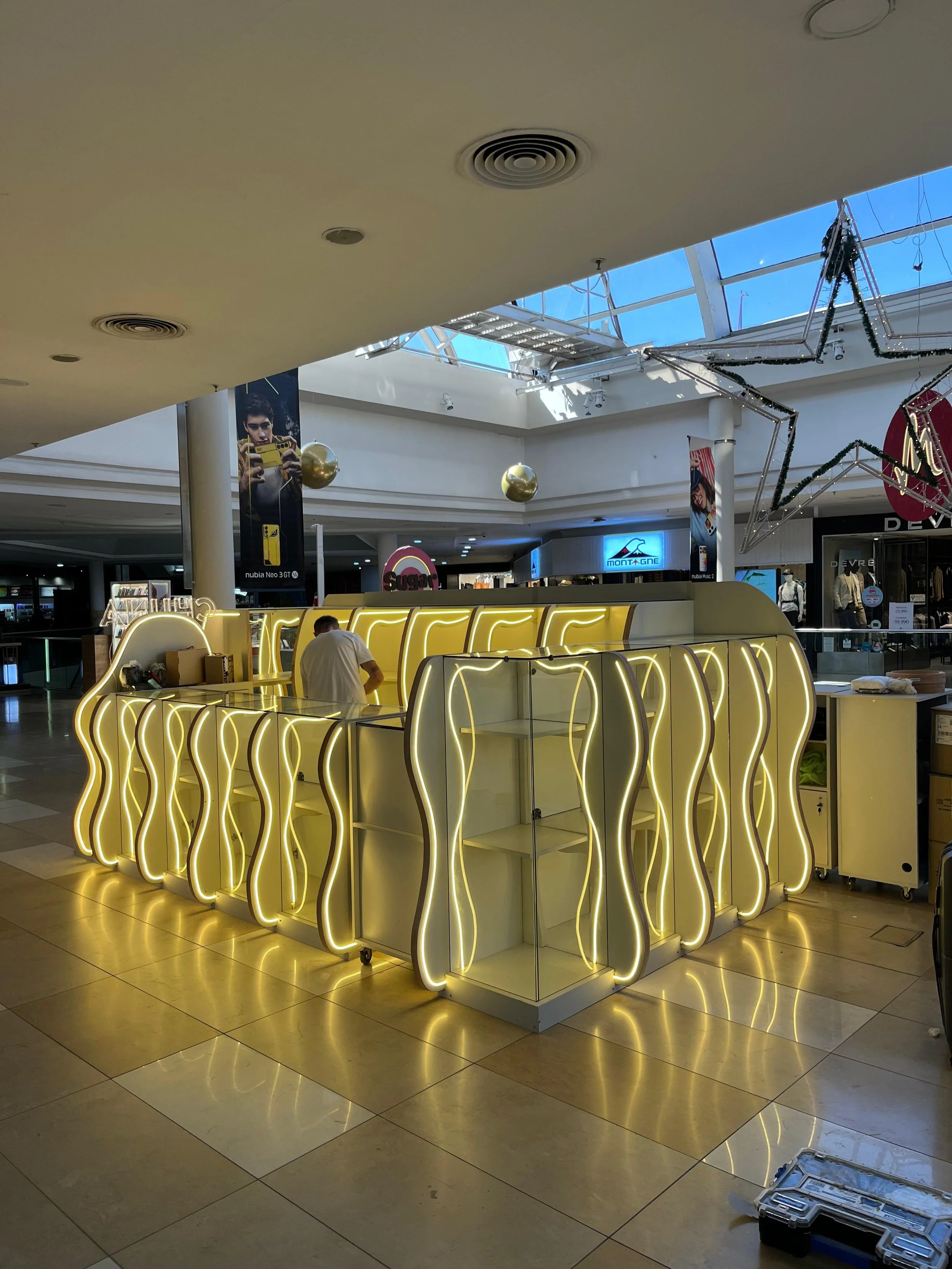 Kiosco de dulces y postres iluminado con luces blancas en un centro comercial, con diseños ondulados en la estructura y una persona atendiendo.