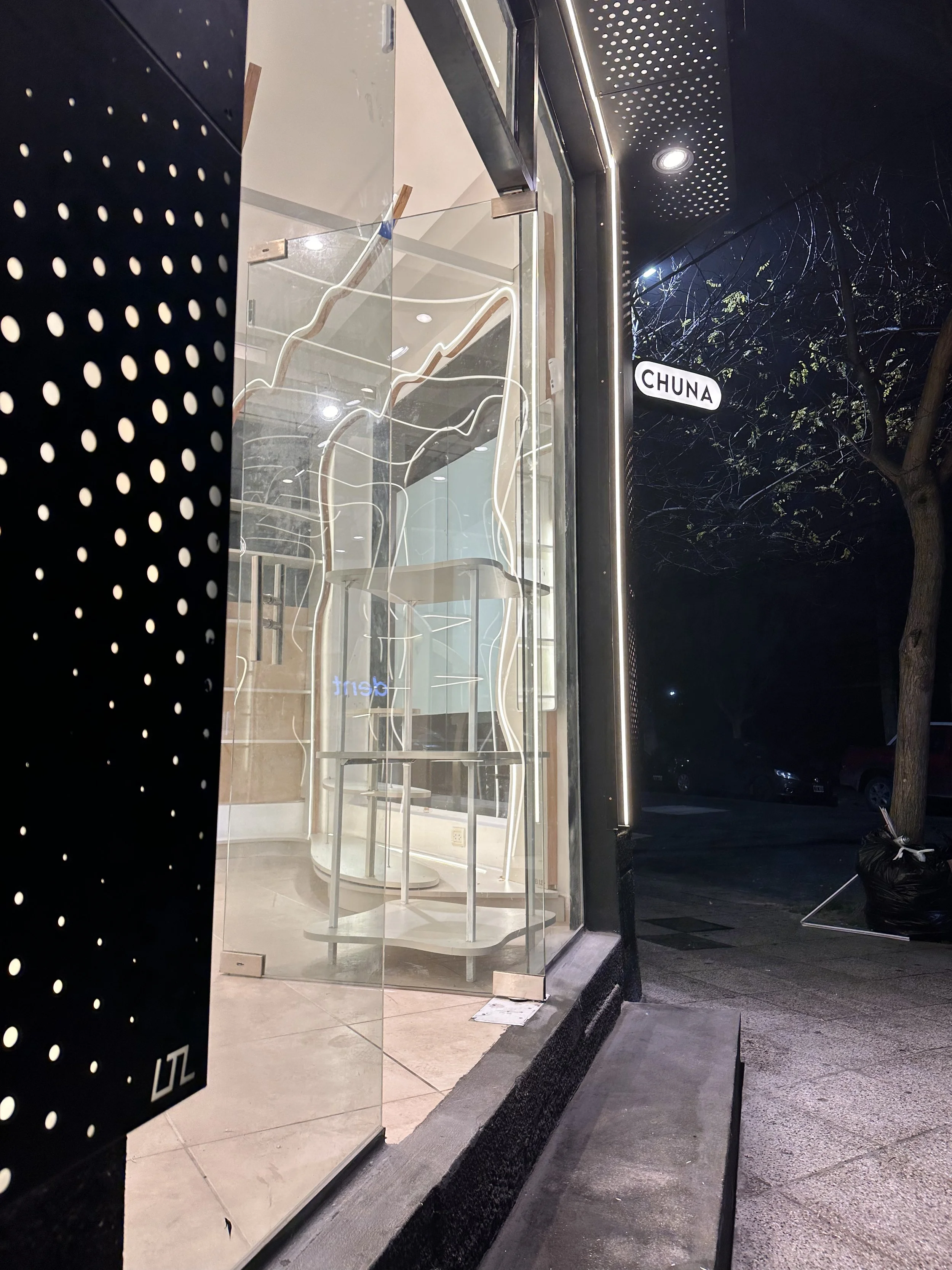 Vista nocturna de la fachada de un negocio con una pared de vidrio, interior con iluminación moderna y mobiliario blanco, indicando la calle 'Chuna' en un letrero en el exterior.