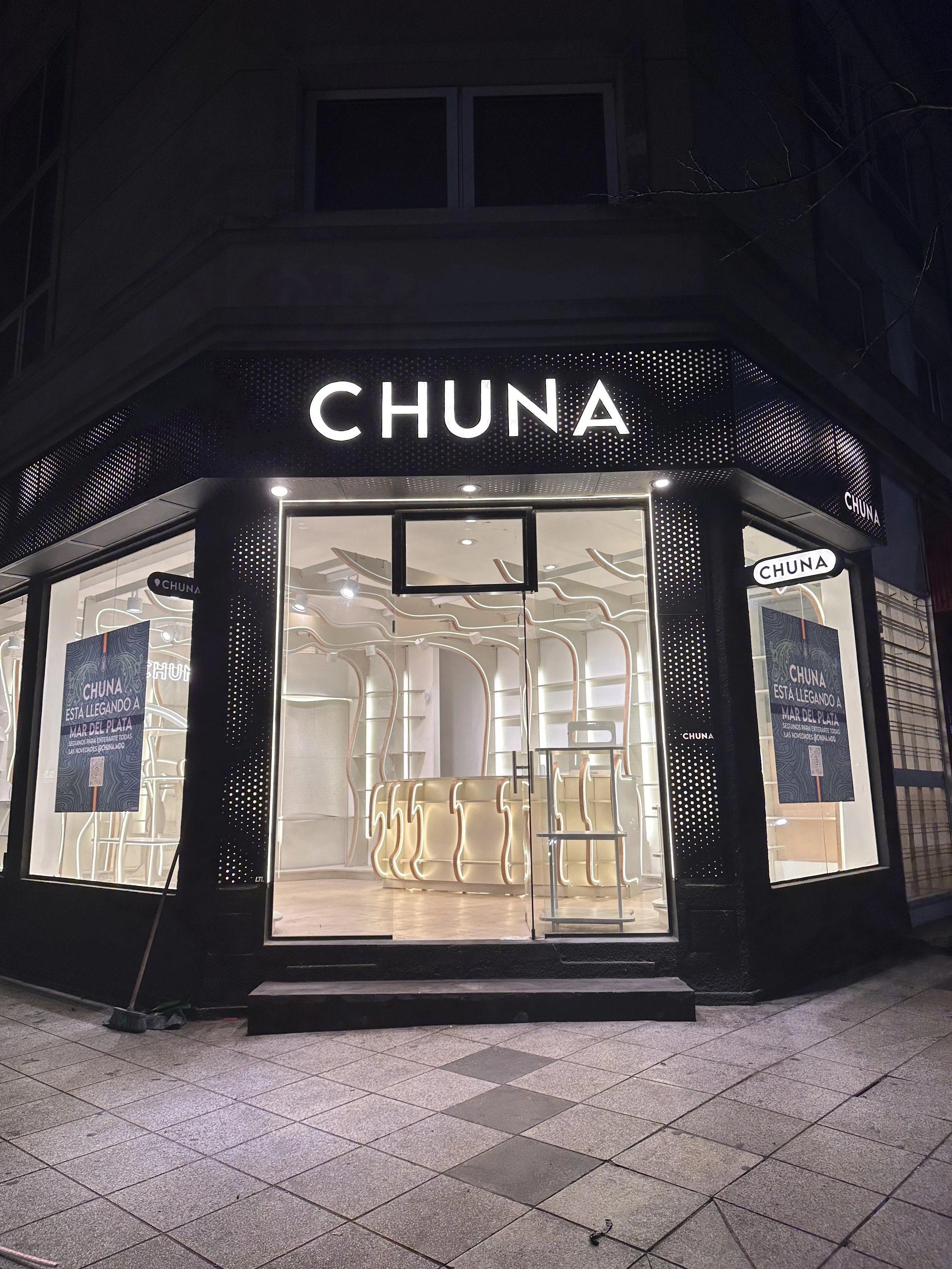 Vista nocturna de una tienda llamada 'CHUNA' con iluminación moderna en la fachada y en el interior.