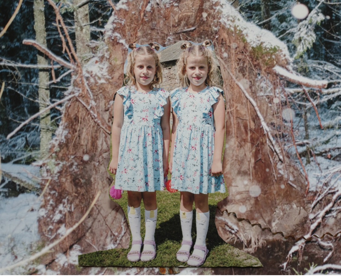Two young girls with curly hair wearing matching blue and white floral dresses, white knee-high socks, and pink sandals, standing outdoors on grass in front of a large tree trunk with snow on the branches, with a forest background.