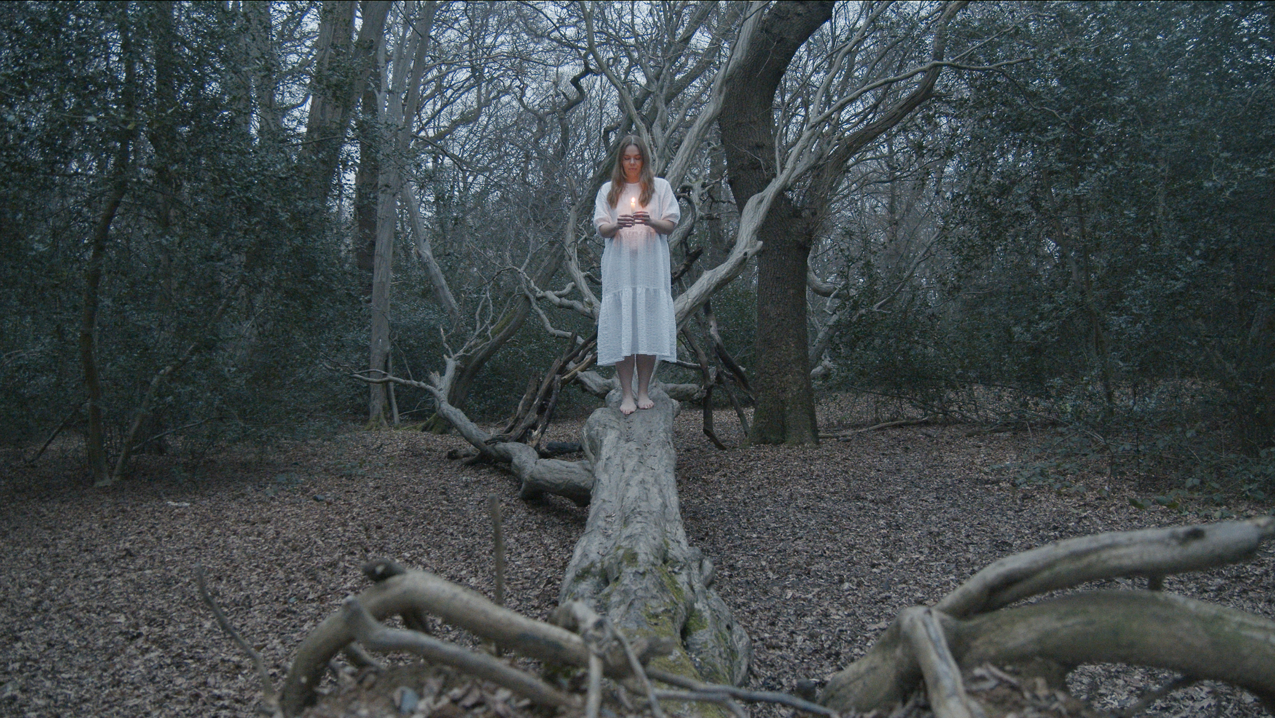 A woman in a white dress standing barefoot on a fallen log in a dark, wooded forest, holding a lit candle.