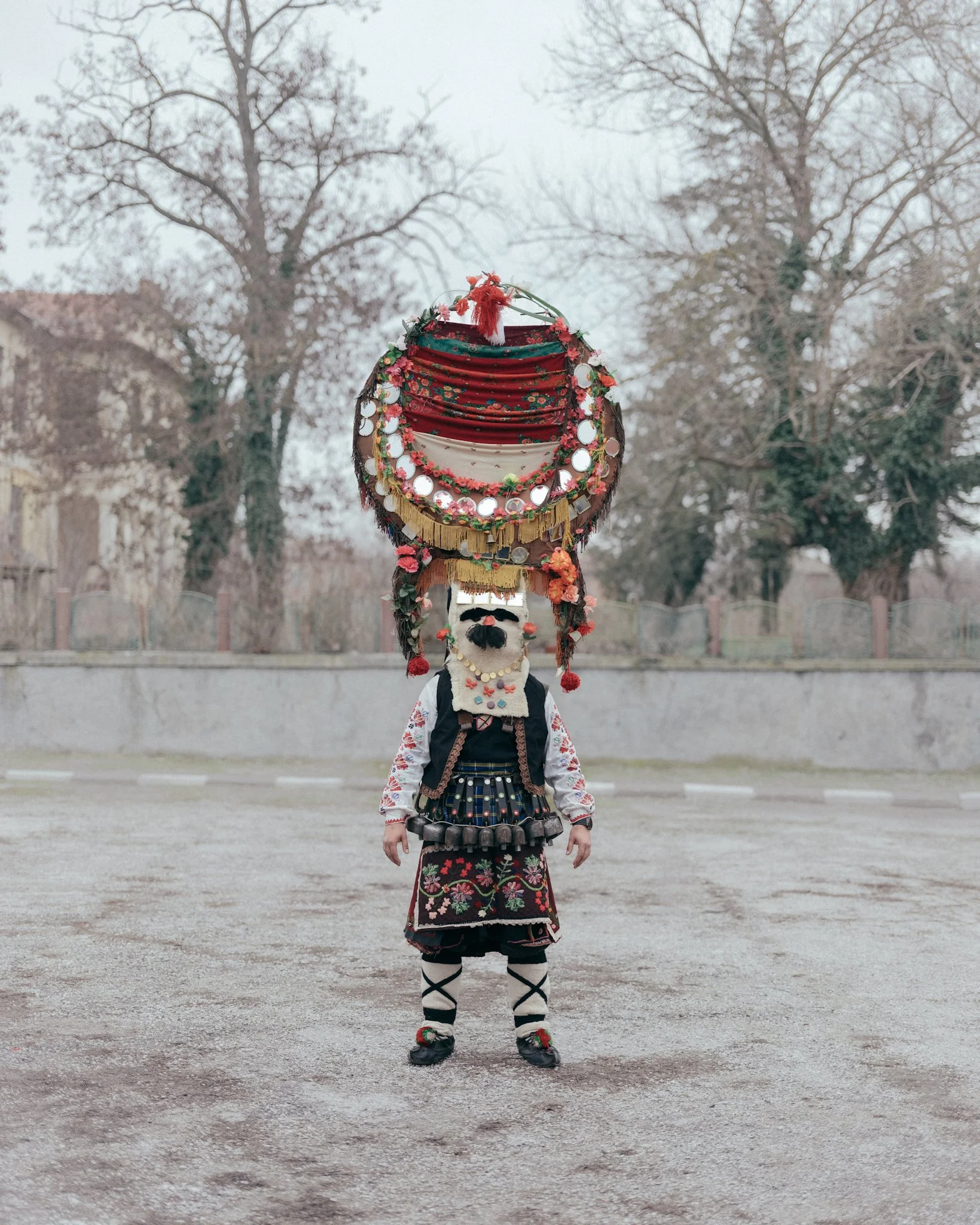 Person dressed in traditional Polish folk costume, holding a large, decorated tambourine-like object on their head, standing outdoors on a gravel surface with leafless trees and a concrete wall in the background.
