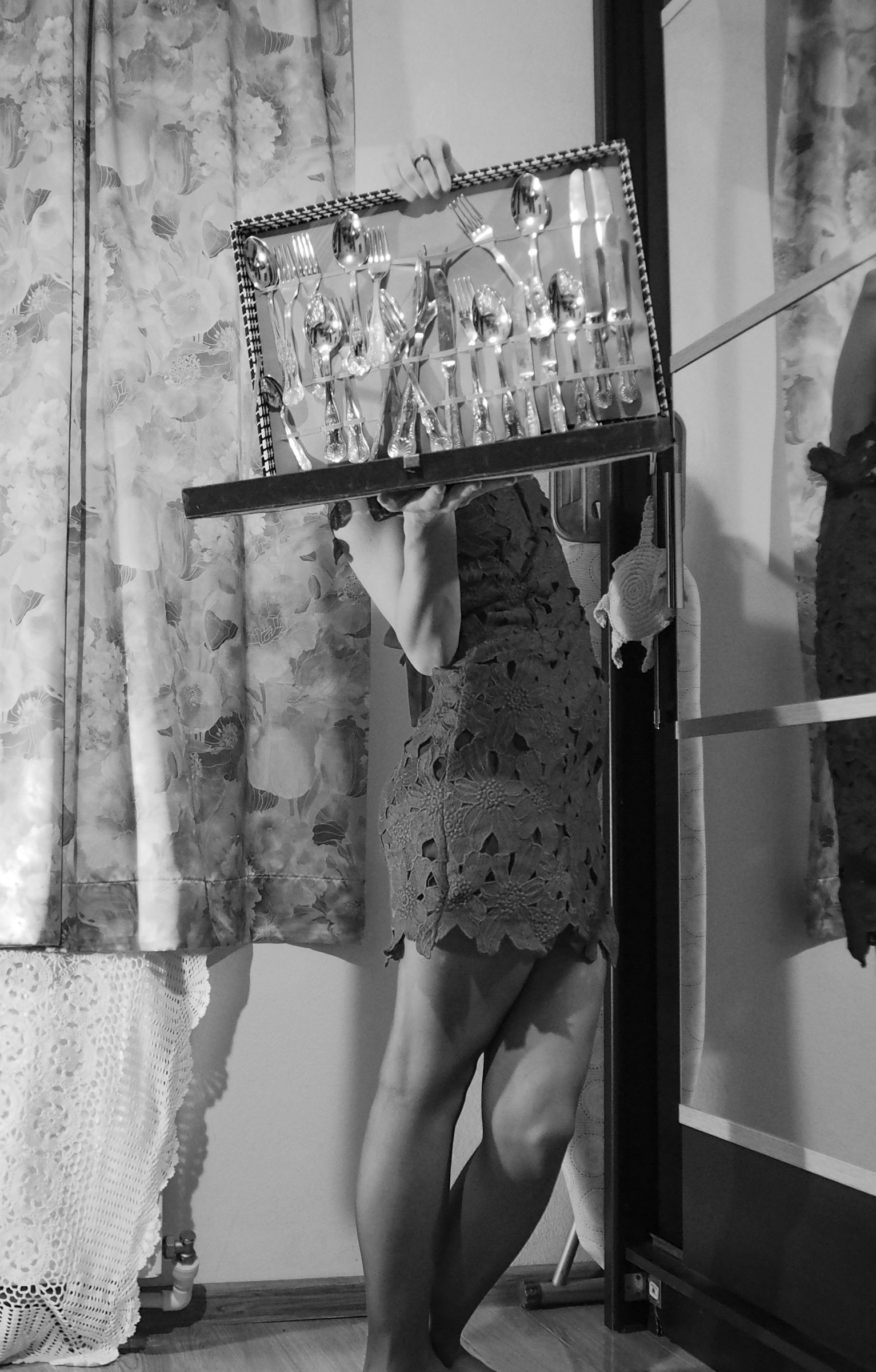 A person in a lace dress holds a tray with an assortment of silverware, including forks, spoons, and knives. The image is in black and white, with floral curtains in the background.