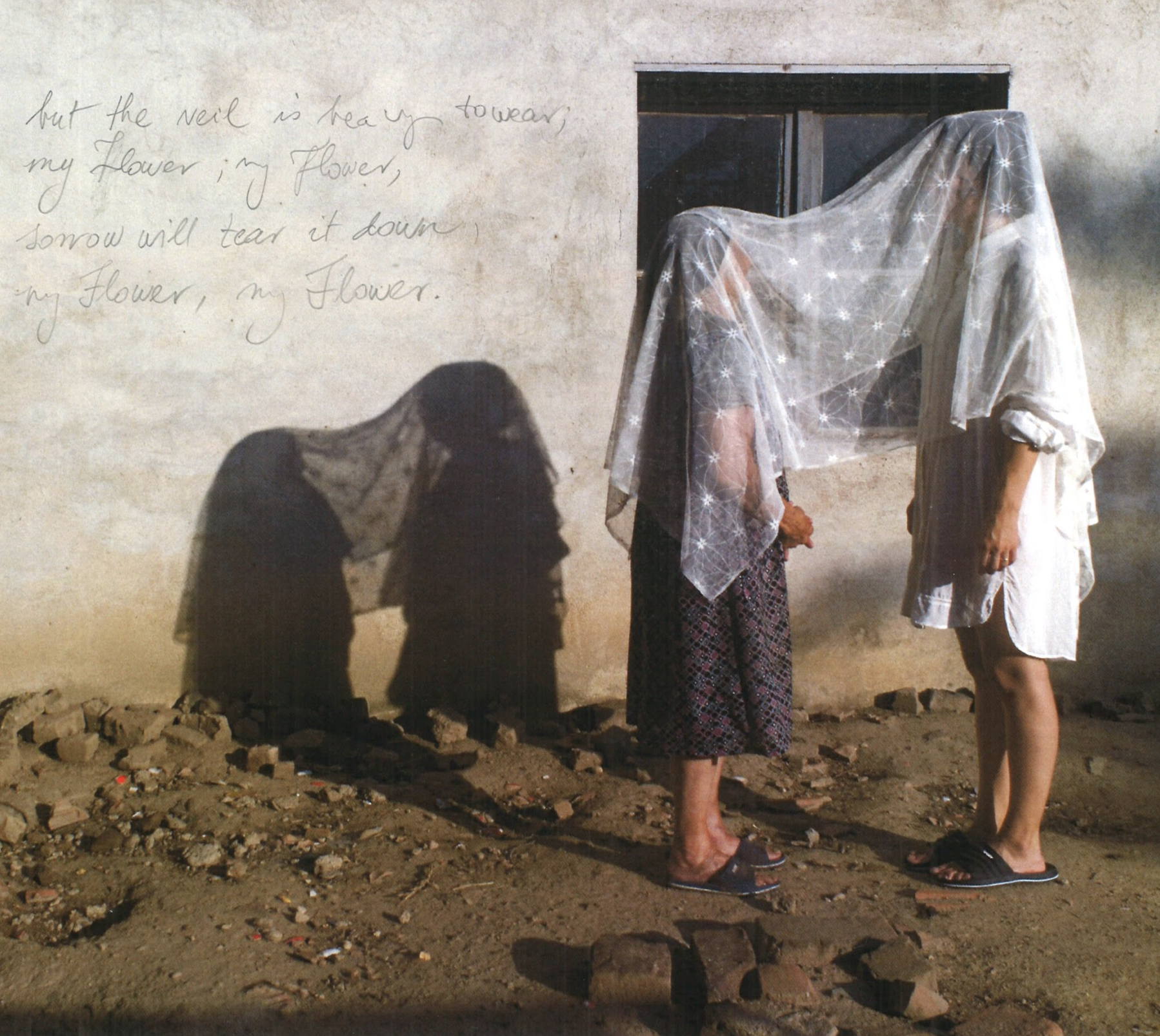 Two women standing outside a building, with a piece of sheer fabric draped over their heads, casting shadows on the wall behind them. The wall has handwritten text that reads, 'but the veil is heavy toward my flower, my flower, sorrow will tear it down, my flower, my flower.'