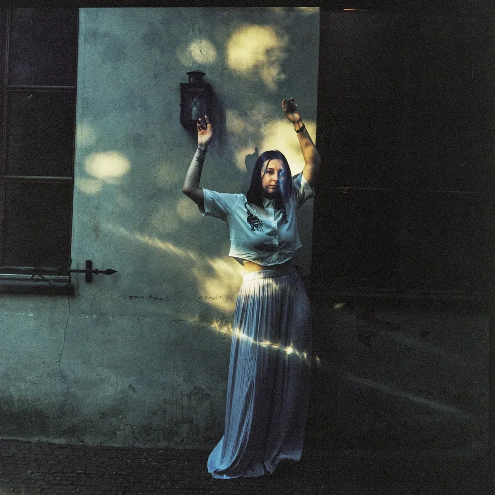 A woman with dark hair standing against a wall, with her hands raised upward. She is wearing a light-colored top and long, wide-leg pants. There is a projection of clouds and a moon on the wall behind her, giving a surreal or artistic effect.