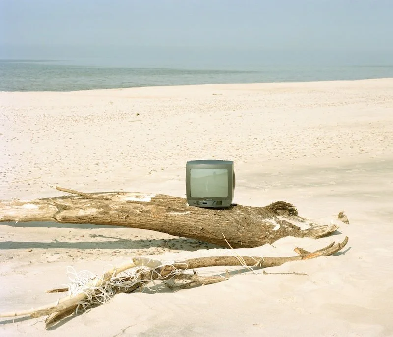 Old television placed on a large piece of driftwood on a sandy beach with the ocean in the background.