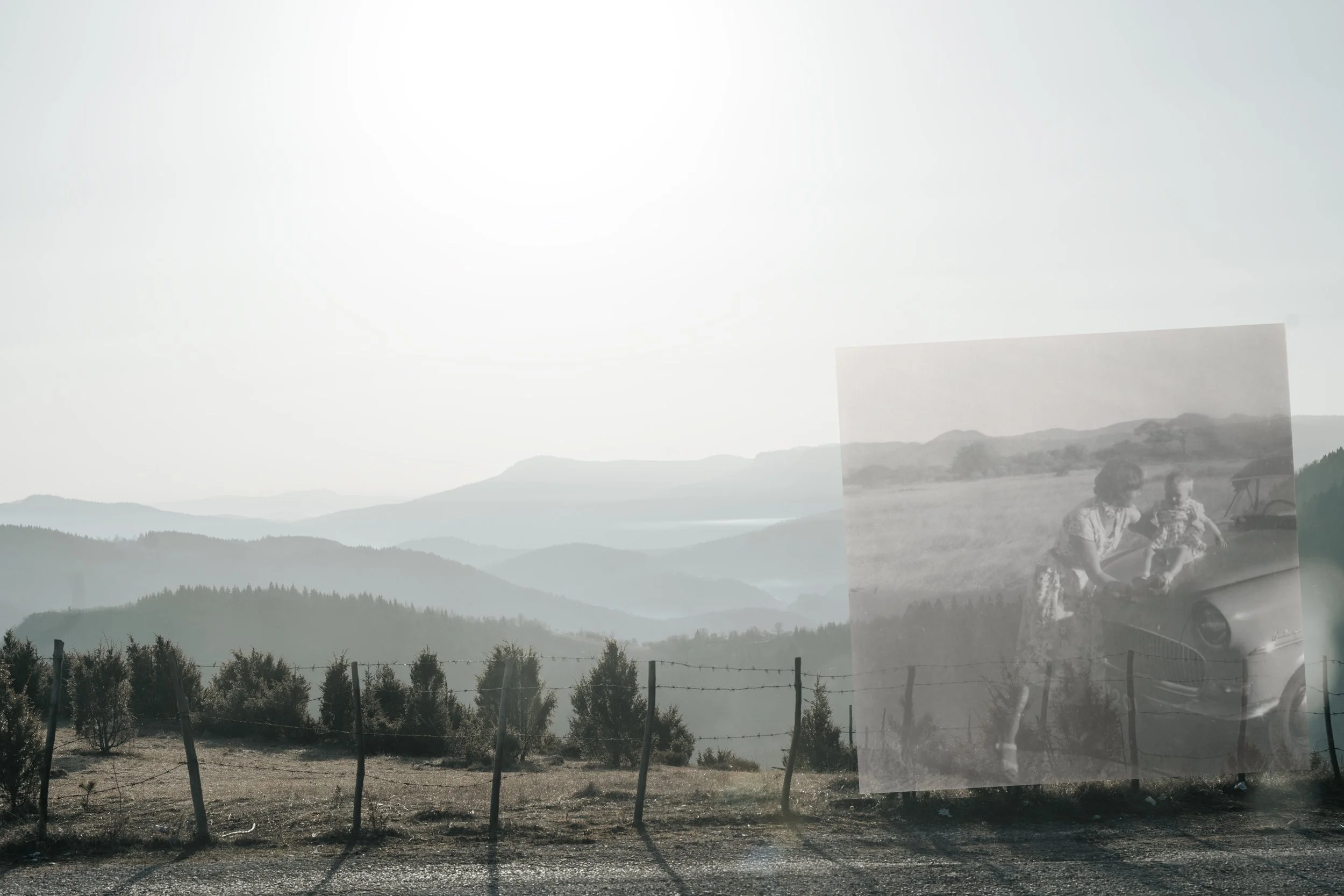 A foggy landscape of rolling hills and trees with a barbed wire fence in the foreground, and a superimposed black-and-white photo of a woman and a small child near a car.