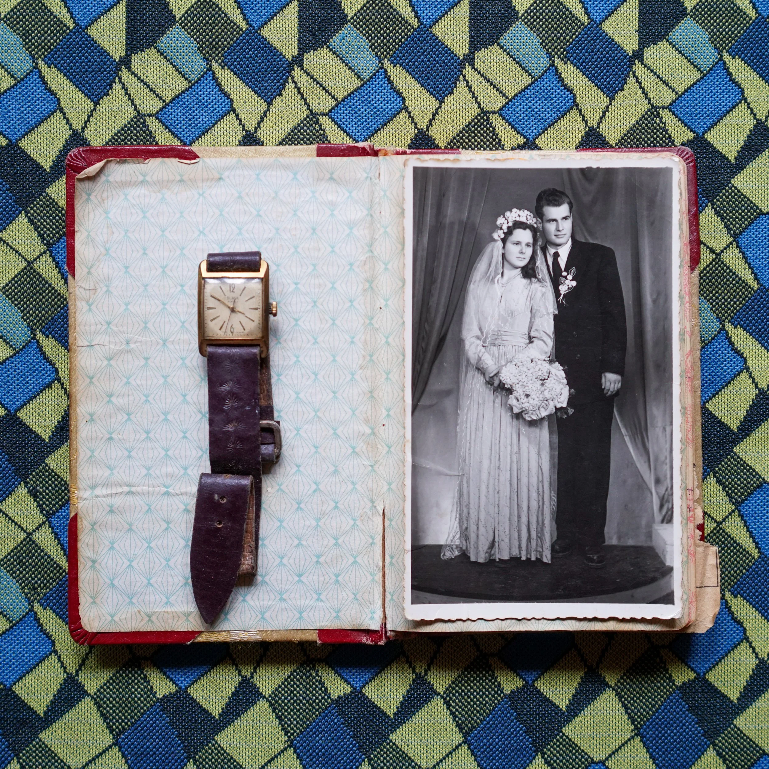 Open photo album on a colorful checkered tablecloth, showing a vintage wristwatch with a leather strap on the left page and a black-and-white wedding photo of a bride and groom on the right page.