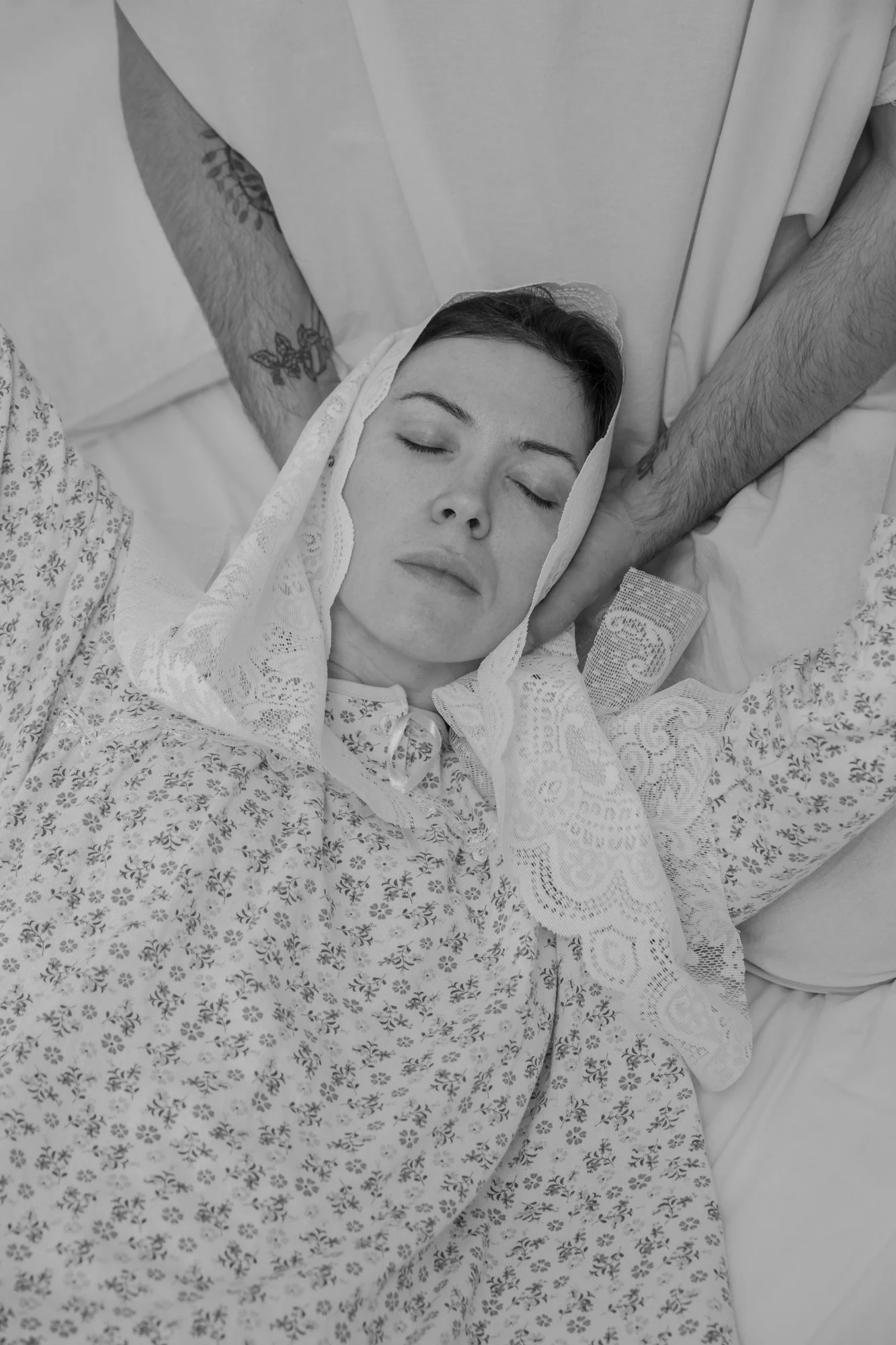 A woman lying in a hospital bed with eyes closed, wearing a patterned hospital gown and a lace veil, with a person supporting her head with their hand.