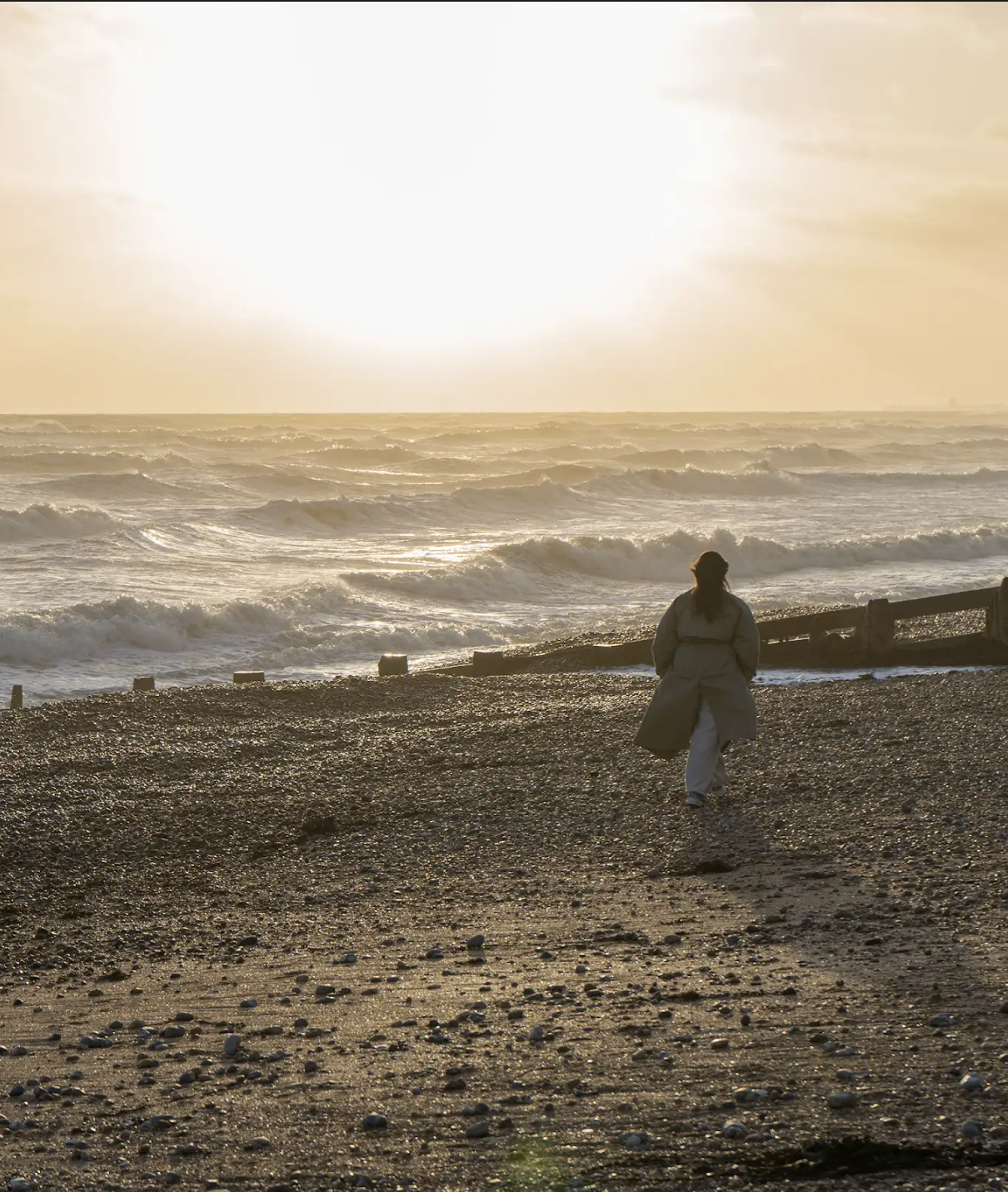 A person walking on a pebbled beach at sunset, overlooking the ocean with waves crashing against the shore.