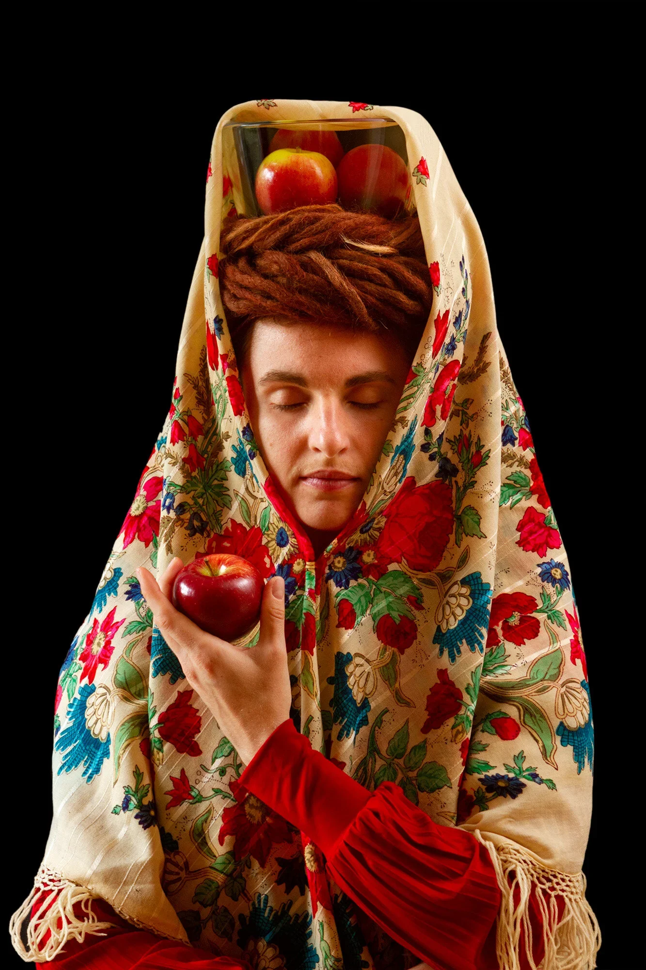 A woman with dreadlocks, eyes closed, holding an apple in her hand, draped in a floral-patterned shawl with a glass of apple juice and two apples on her head on a black background.