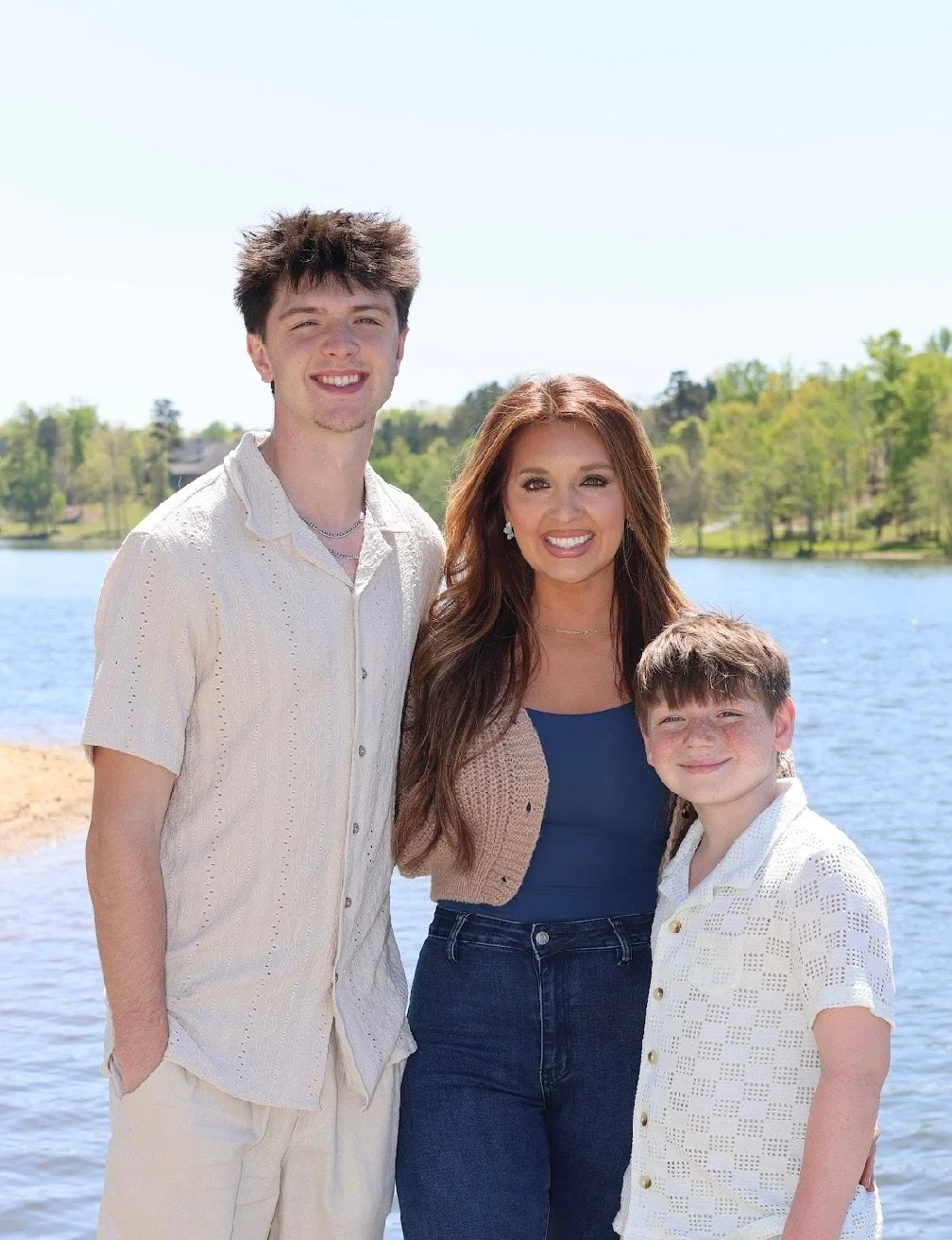 A woman and two young men standing by a lake on a sunny day, smiling at the camera.