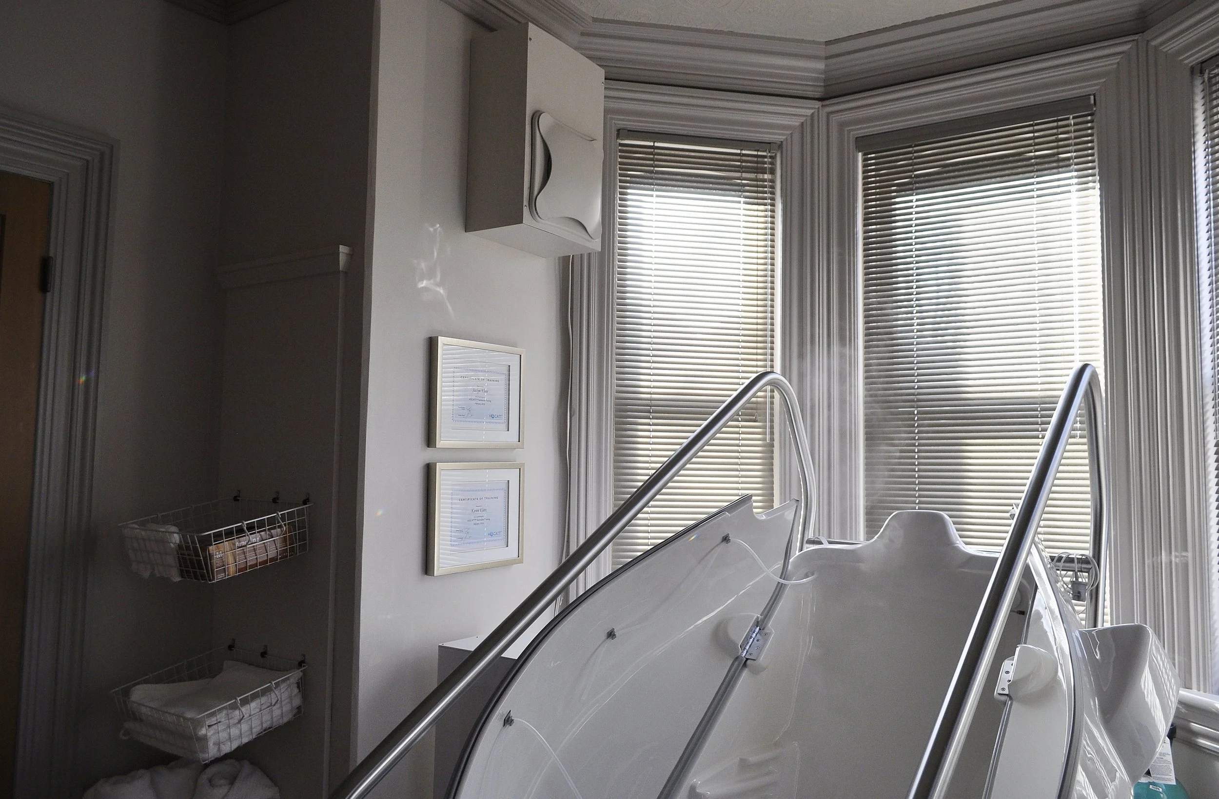 Insider view of a medical or wellness spa with a reclining spa chair, large windows with blinds, framed certificates on the wall, and storage baskets with towels.