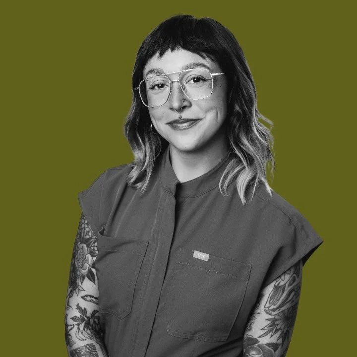 A woman with shoulder-length, wavy hair wearing glasses and a tattoo on her arm, smiling in front of a solid olive-green background.
