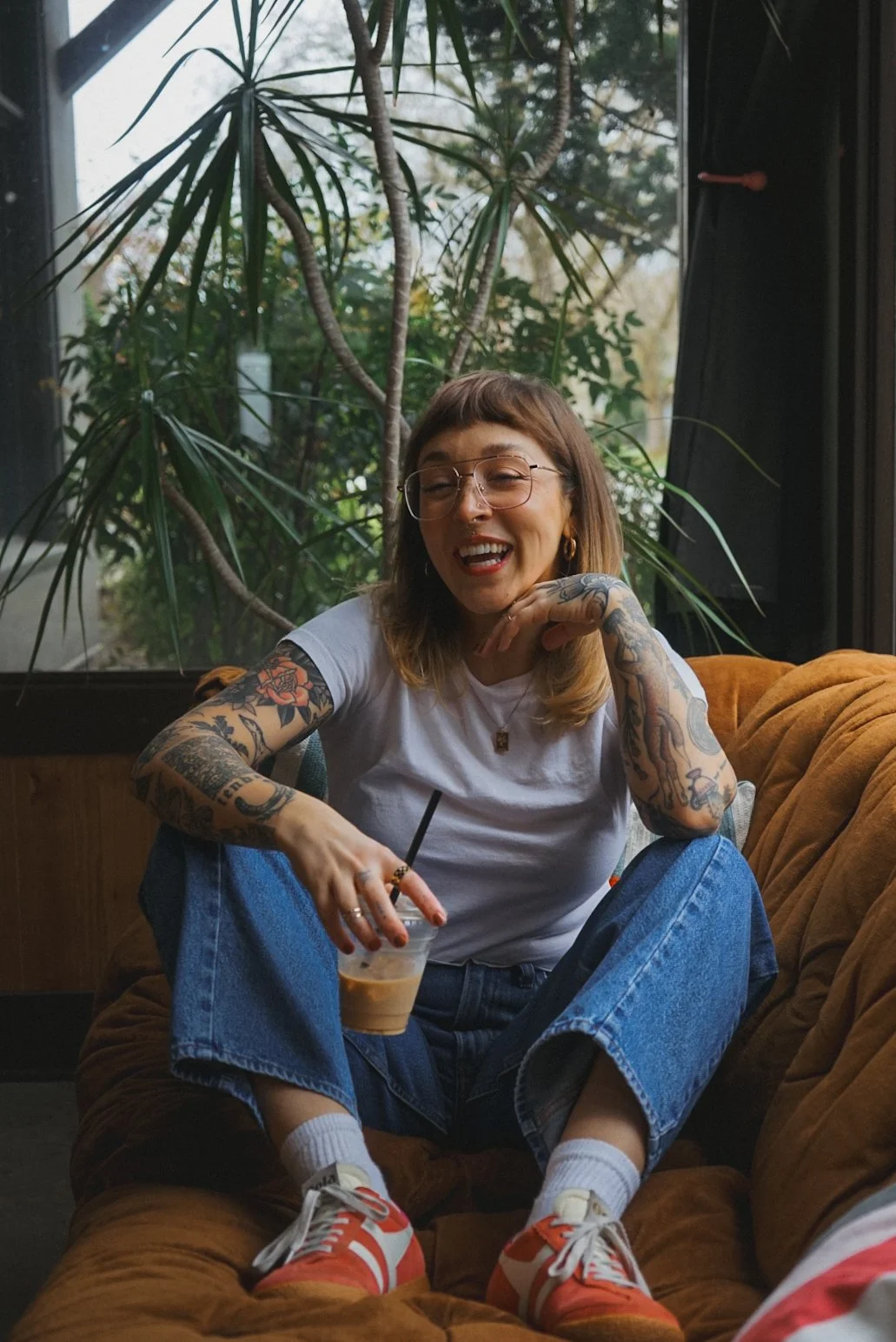 A woman with tattoos, glasses, and shoulder-length hair sitting on a mustard-colored sofa, smiling, holding an iced coffee, in front of a large window with greenery outside.