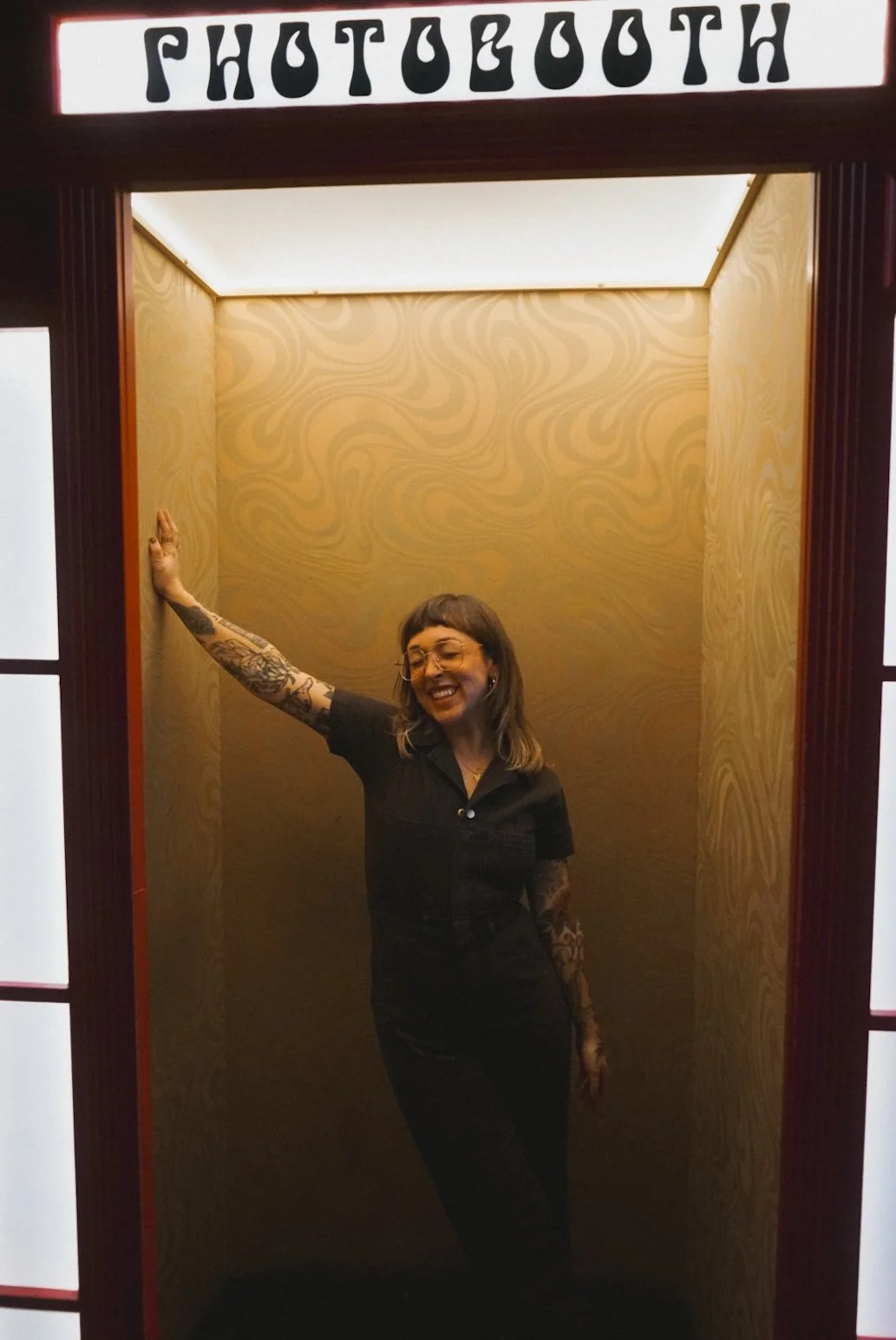 A woman with tattoos on her arms, wearing glasses and a black outfit, smiling inside a photo booth with a gold patterned background.
