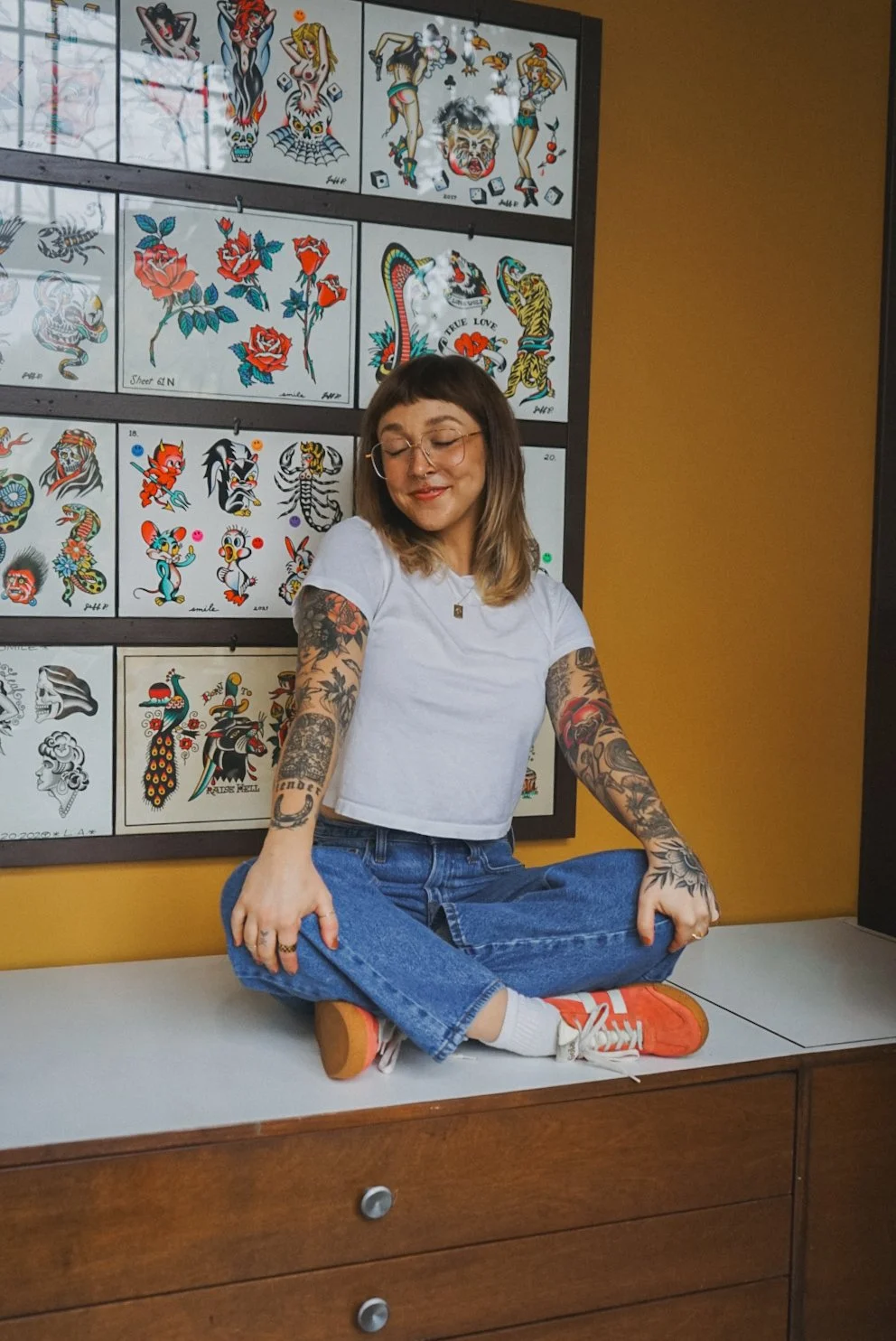 A woman with tattoos sitting cross-legged on a wooden table, in front of a wall with tattoo flash art.