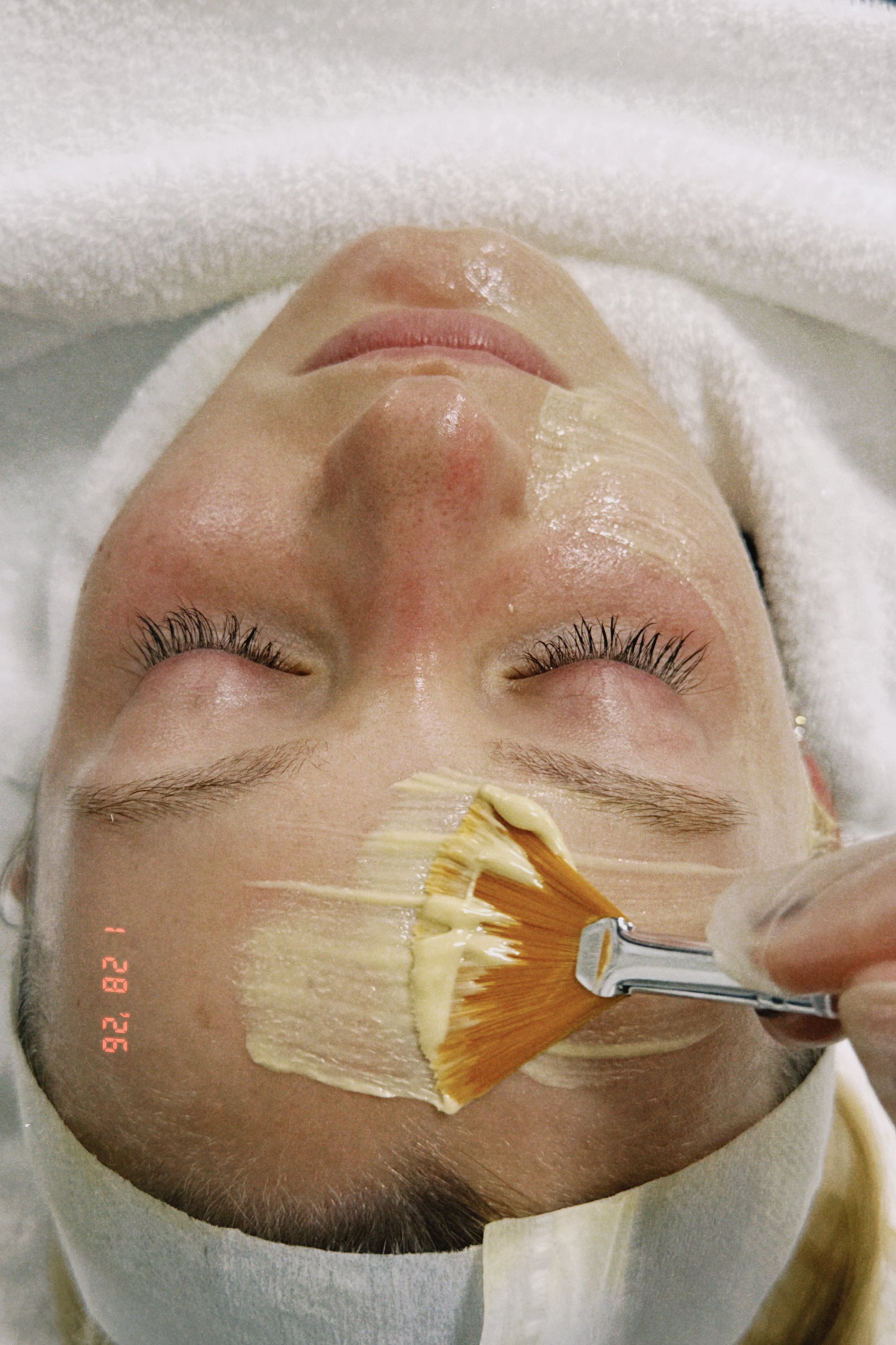 A woman with closed eyes receiving a facial treatment, with a brush applying a mask or cream to her face, lying on a treatment bed covered with a towel.