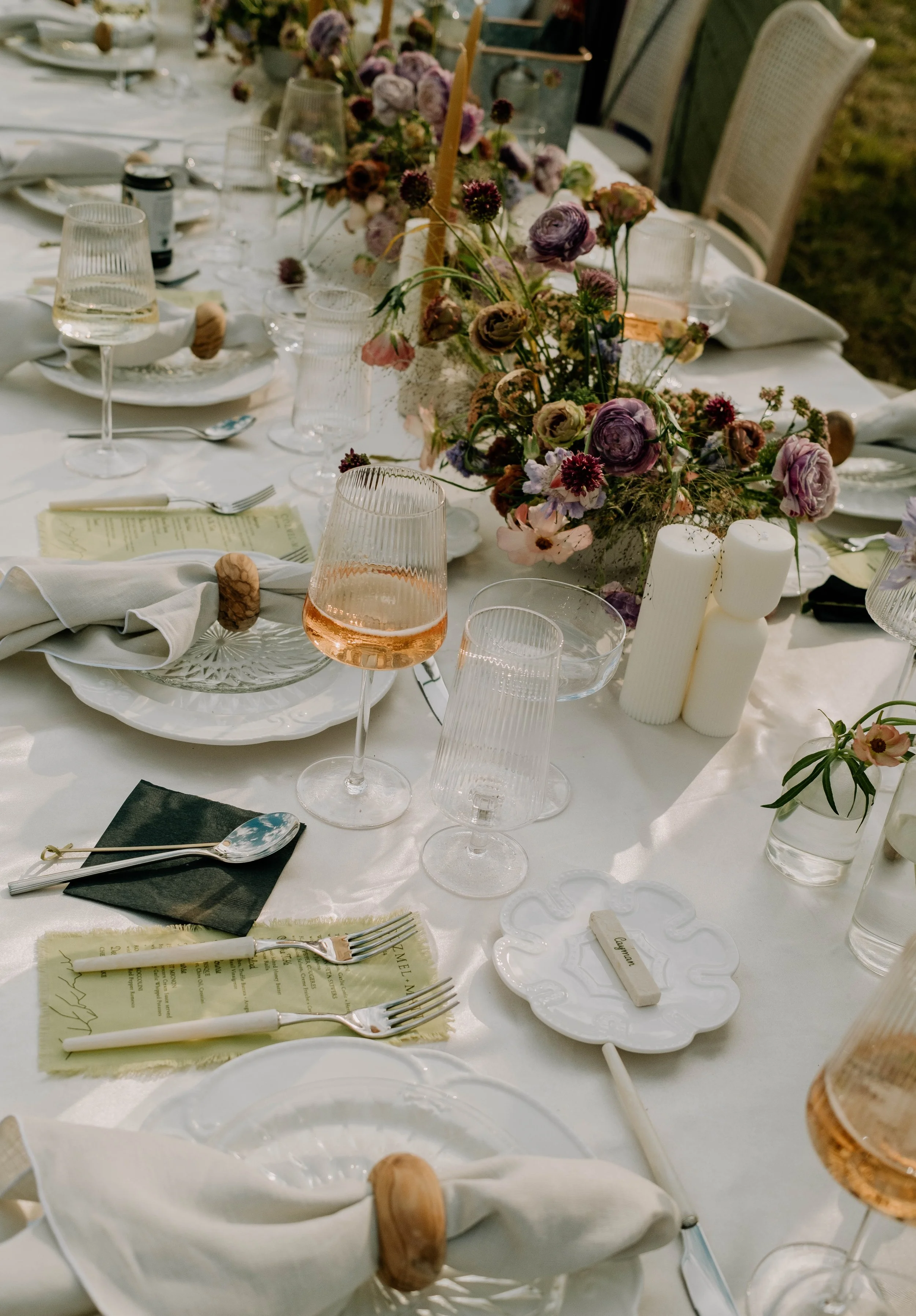 A formal dining table decorated with a floral centerpiece, candles, wine glasses, plates, and utensils set for a special occasion, outdoors at sunset.