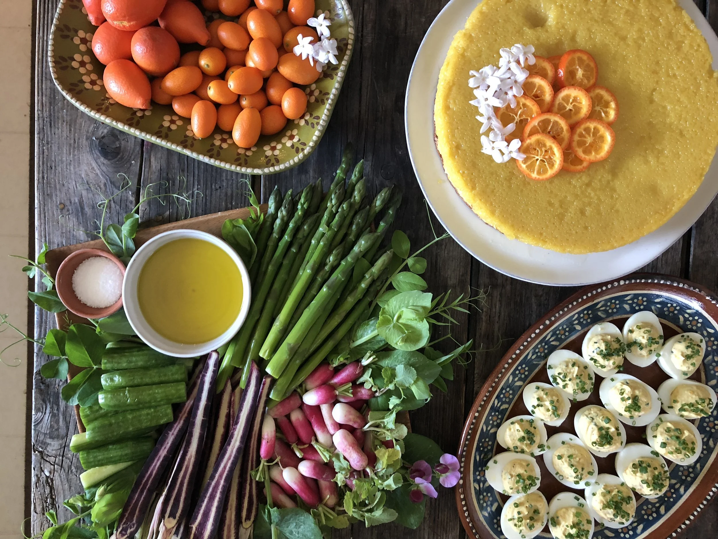 A wooden table with a variety of food items and ingredients including fresh orange cherry tomatoes and kumquat citrus slices on a yellow cake decorated with kumquat slices and small white flowers, cucumber, radish, asparagus, a bowl of olive oil, a small bowl of salt, and a platter of deviled eggs garnished with chives.
