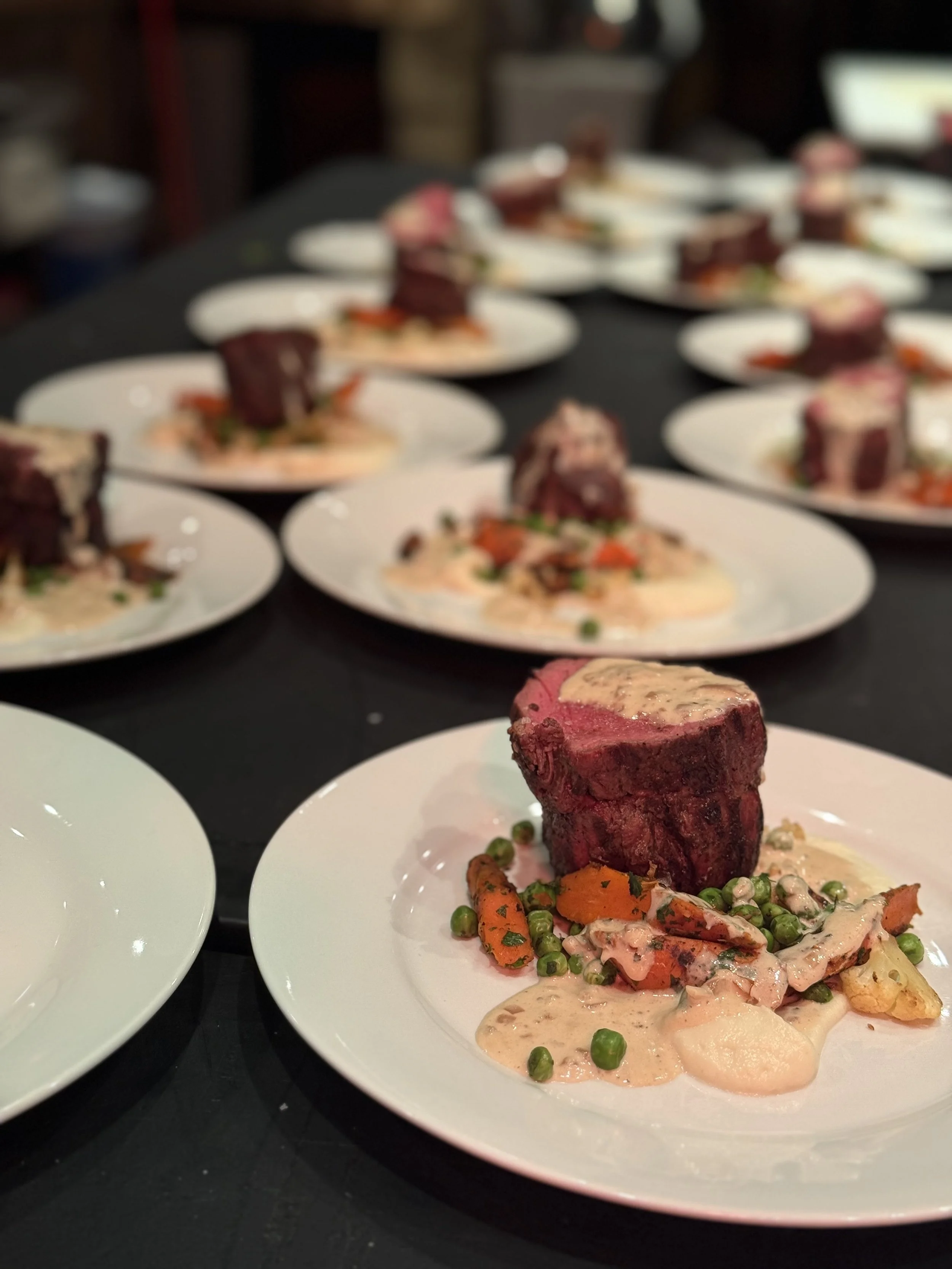 Several plates with cooked beef tenderloin topped with sauce, accompanied by vegetables including carrots, peas, and potatoes, on a dark table.