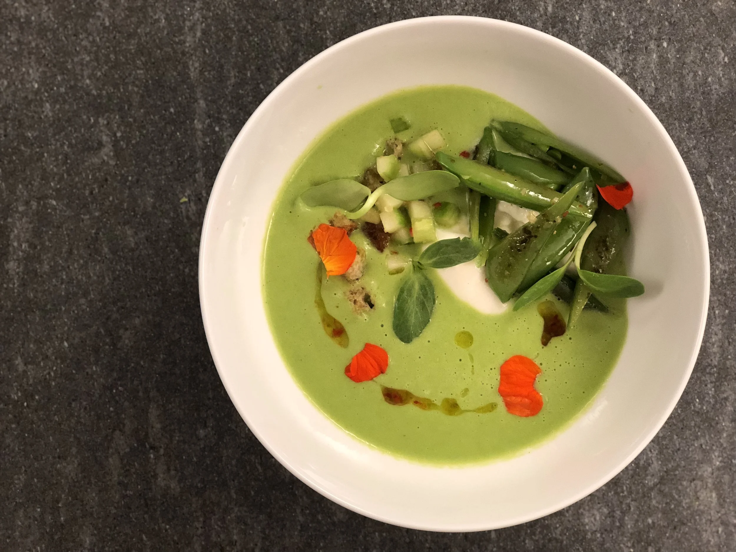 A bowl of green soup garnished with edible flowers, herbs, and vegetables on a dark surface.