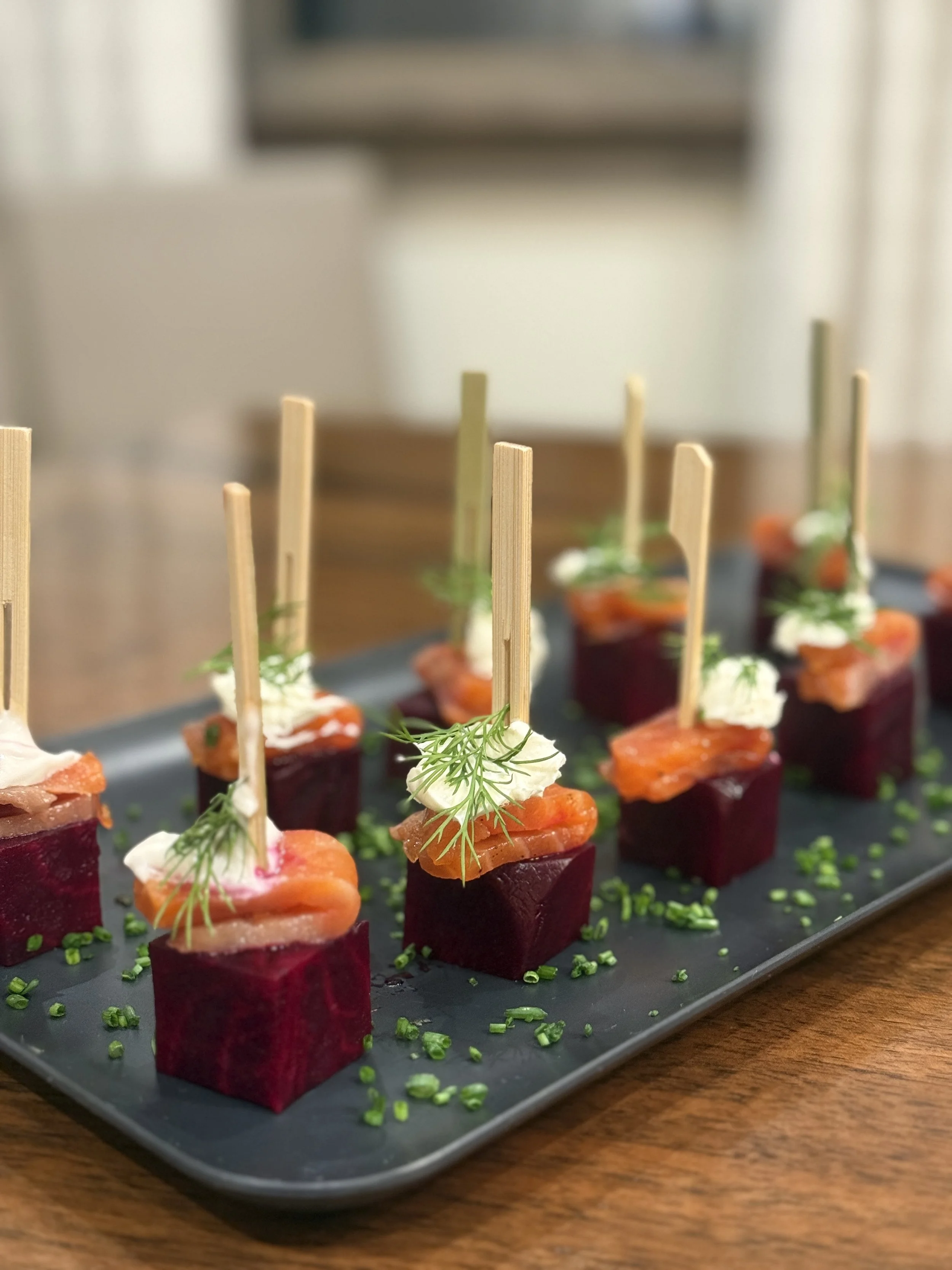 Appetizers on a black rectangular platter, consisting of diced beets topped with smoked salmon, garnished with dill and cream, and skewered with wooden sticks, sprinkled with chopped chives.