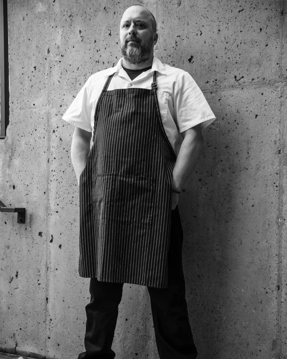 A man with a beard wearing a white shirt and a striped apron standing against a textured concrete wall.