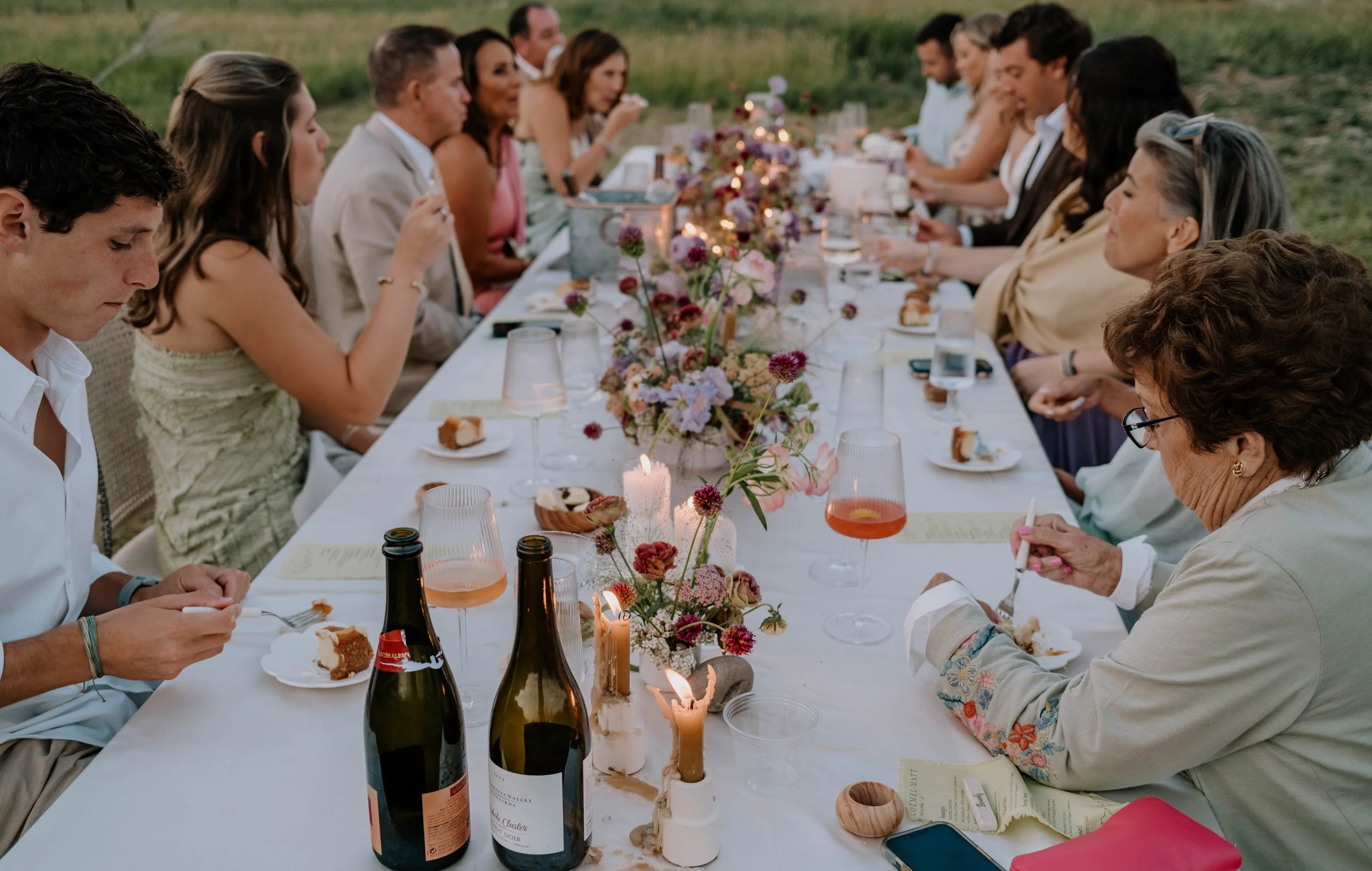 People sitting at a long outdoor dinner table decorated with flowers and candles, enjoying a meal together.