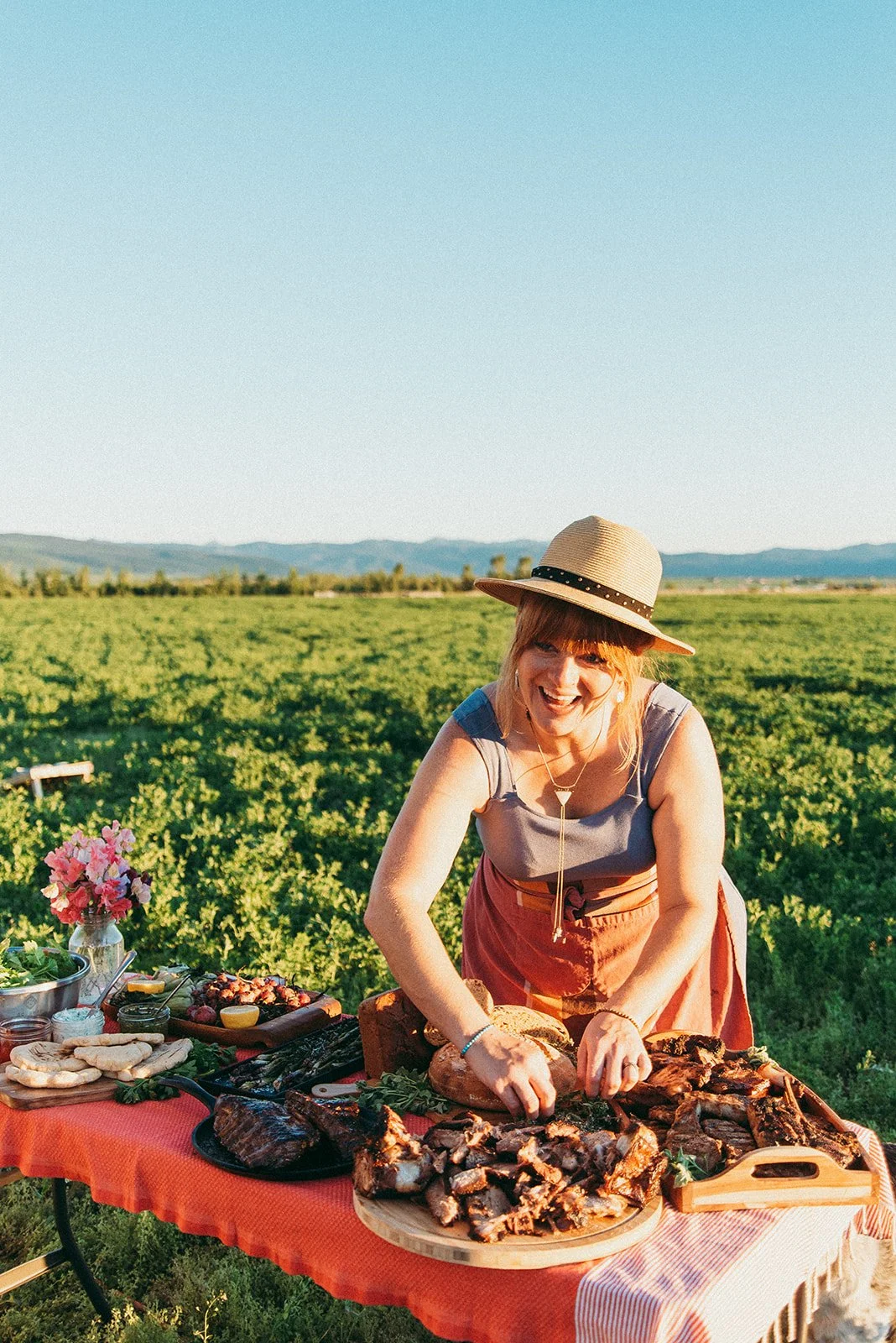 A woman with a straw hat and gray sleeveless top smiling and slicing cooked meat at an outdoor barbecue table in a green field with mountains in the background.